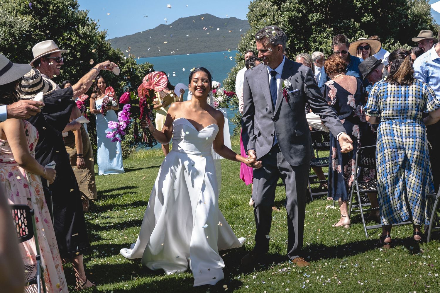 Bride and groom walk through confetti shower outdoors by lakeside, surrounded by cheering wedding guests on sunny day.