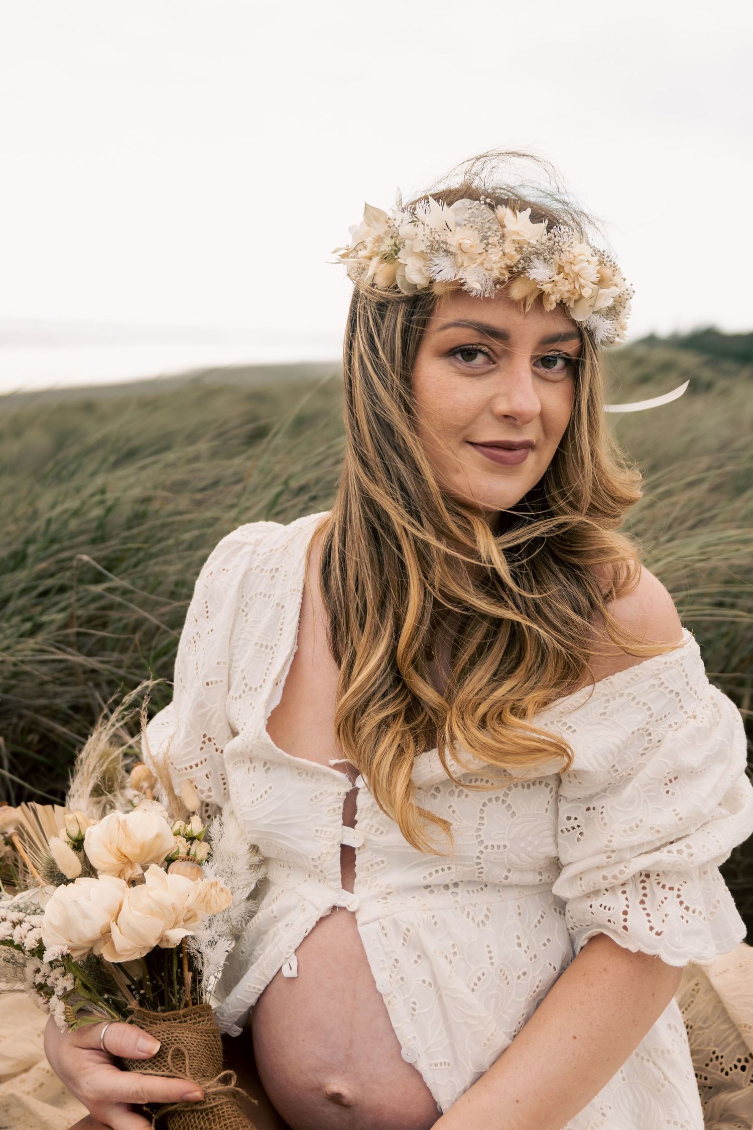 Pregnant woman in white boho dress with floral crown holding bouquet outdoors near grassy dunes.