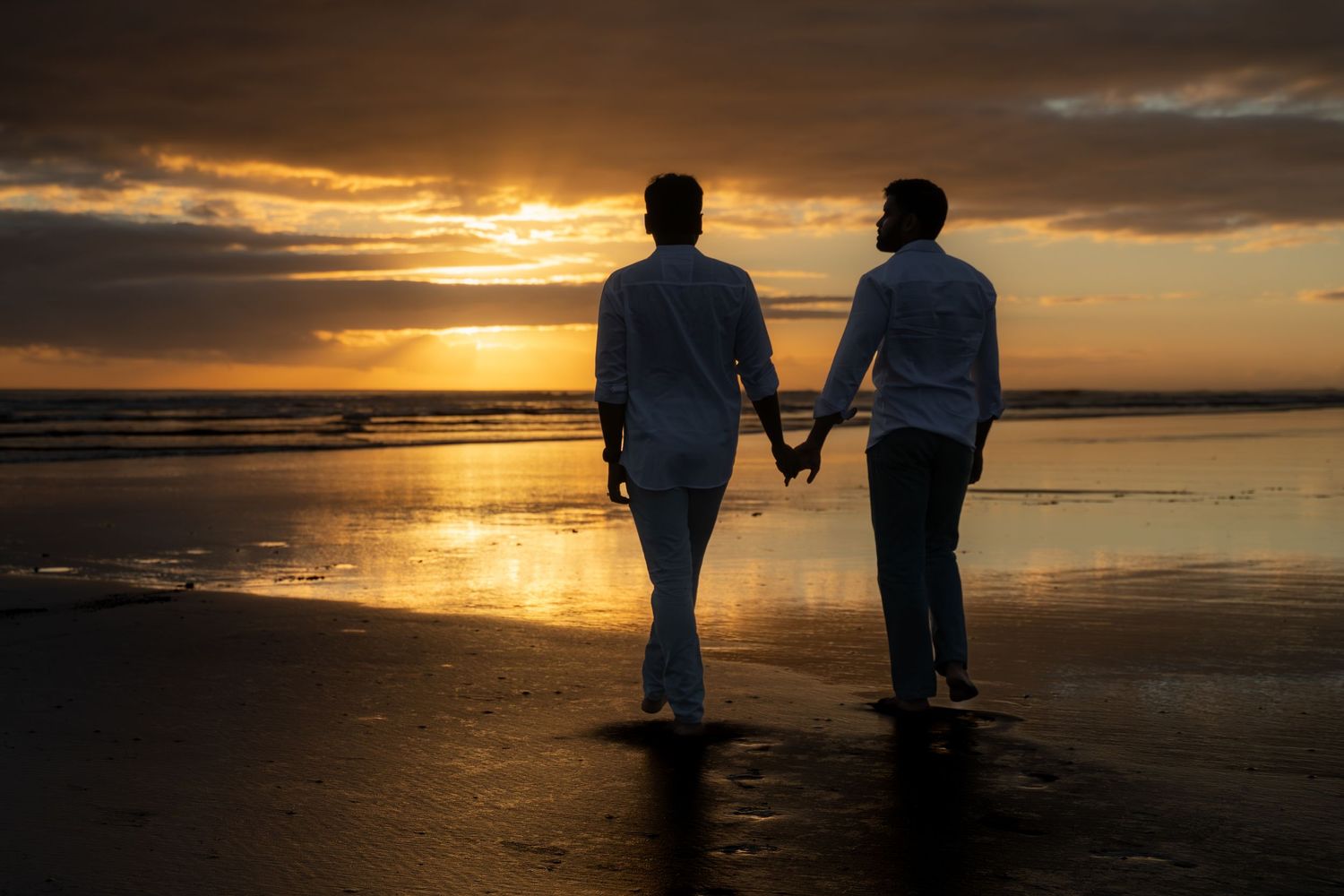 Two people holding hands walking on a beach at sunset, silhouetted against a golden sky.