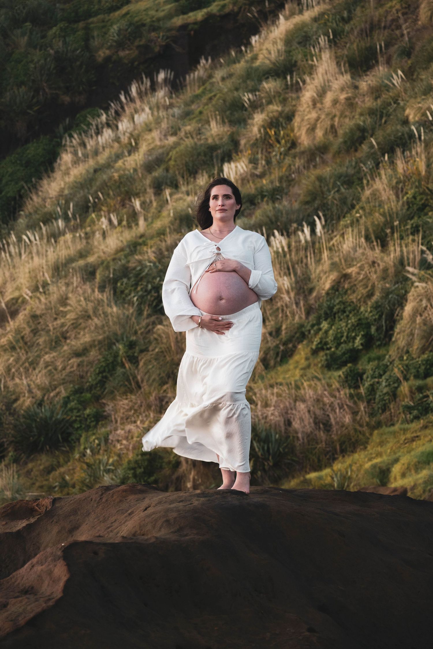 Pregnant woman in white dress holding baby bump outdoors on rocky terrain with green hillside backdrop.