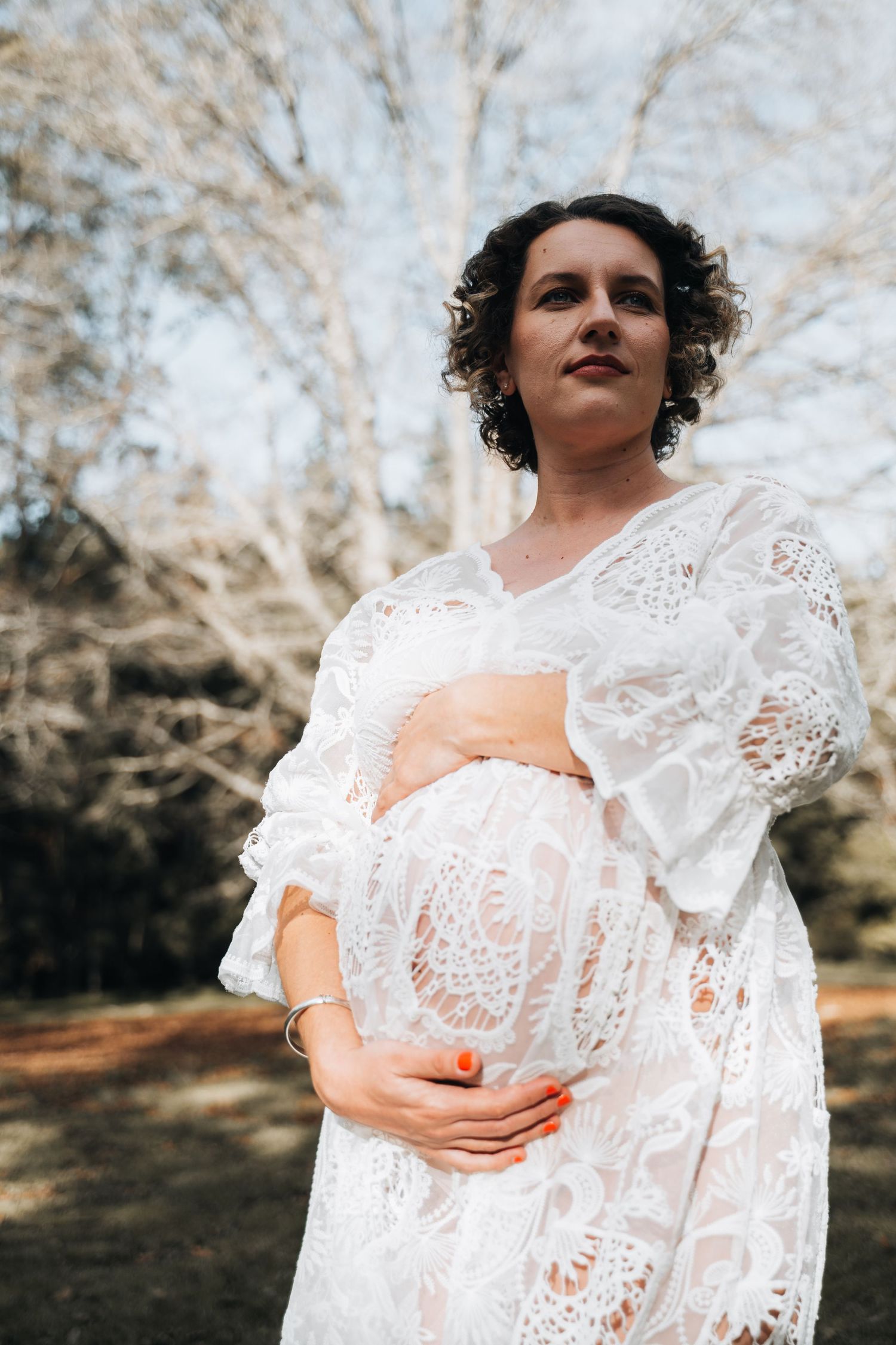 Pregnant woman in white lace dress cradling baby bump outdoors among bare trees.