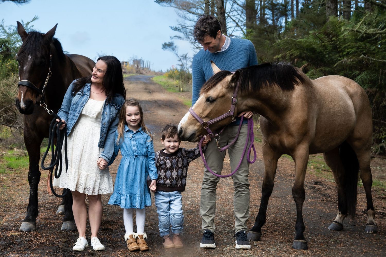 Family with two children posing outdoors with two horses on a forest trail.