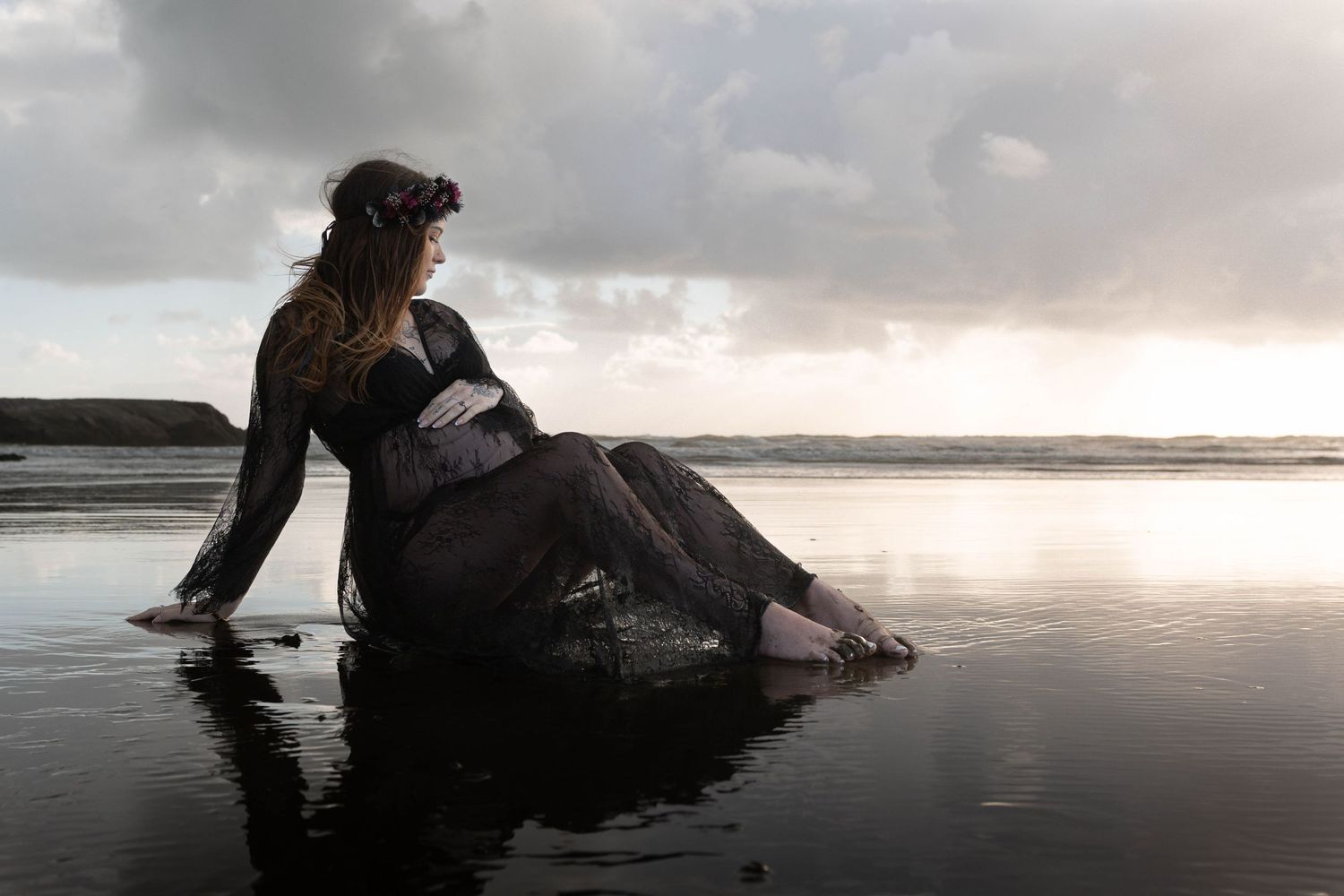 A woman in a dark dress sits on a rock at a reflective beach during a dramatic cloudy sunset.