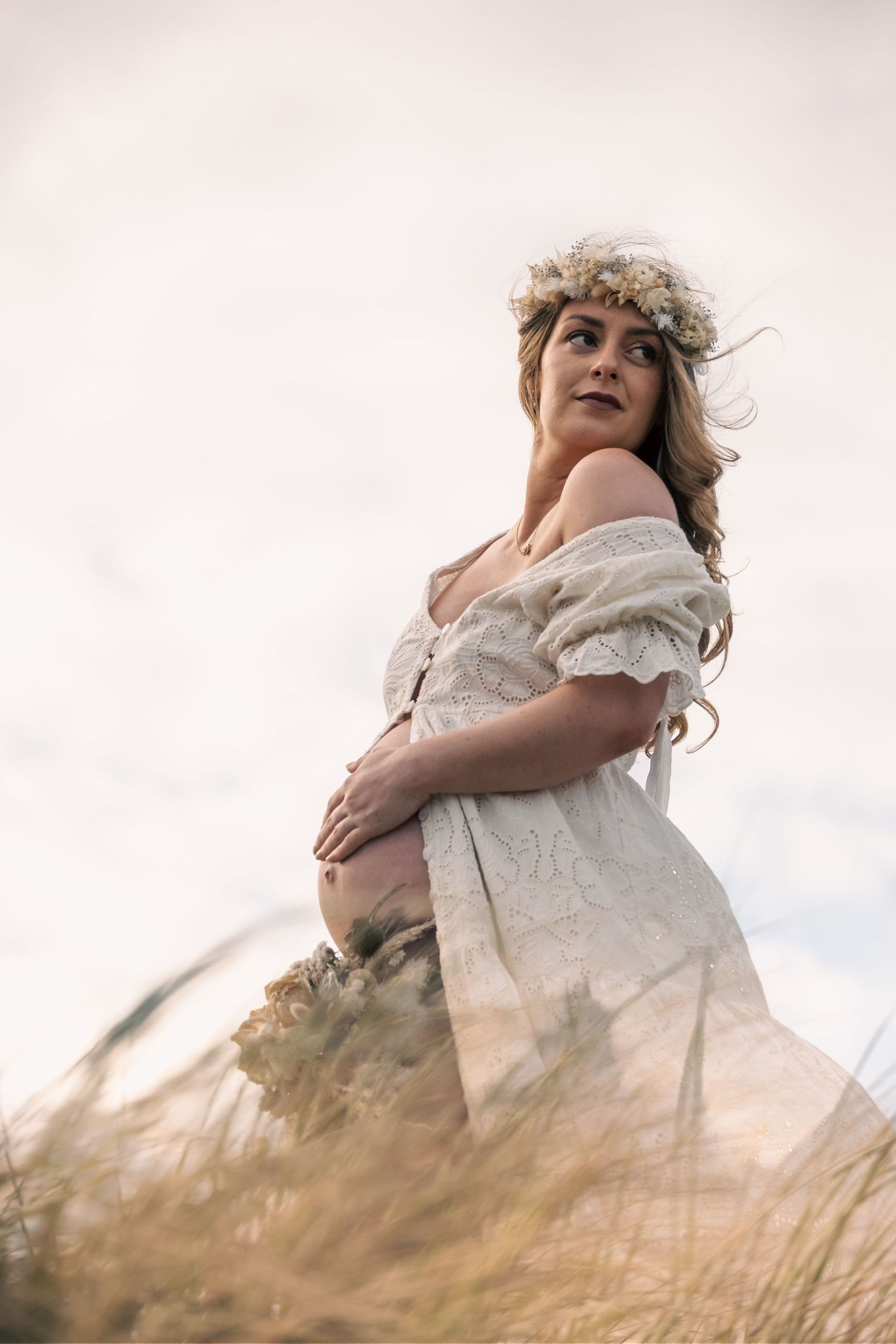 Pregnant woman in white lace off-shoulder dress and floral crown stands in golden grass field under soft sky.