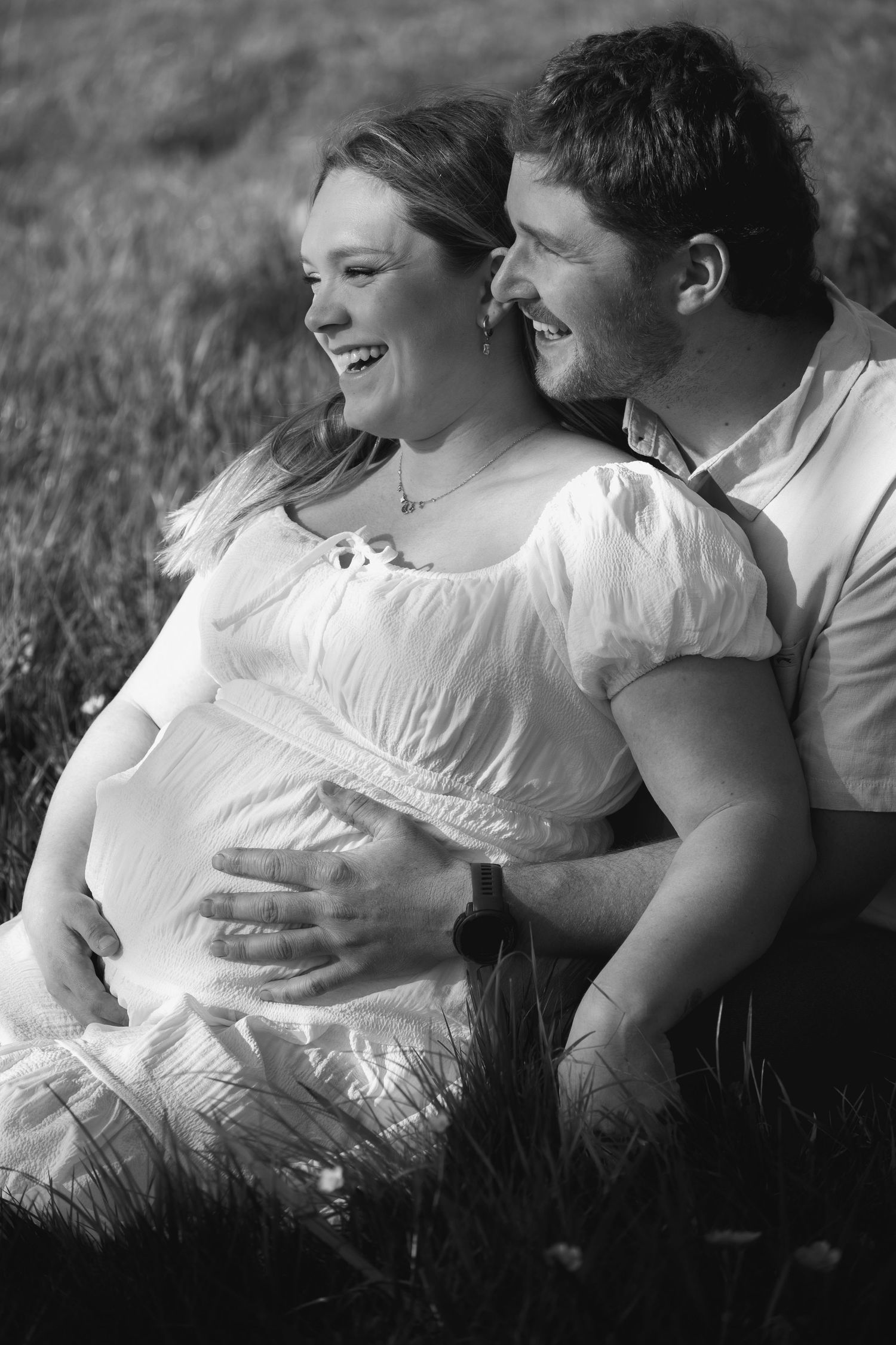 Black and white photo of a laughing pregnant couple sitting in a grassy field, man embracing woman's baby bump.
