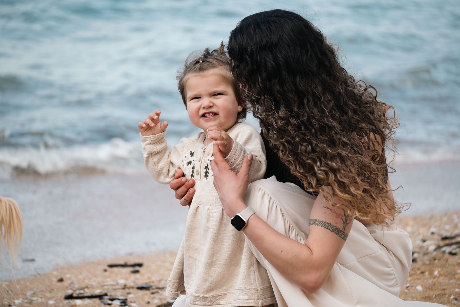 A mother with curly dark hair kisses her smiling toddler daughter on a rocky beach by the ocean.