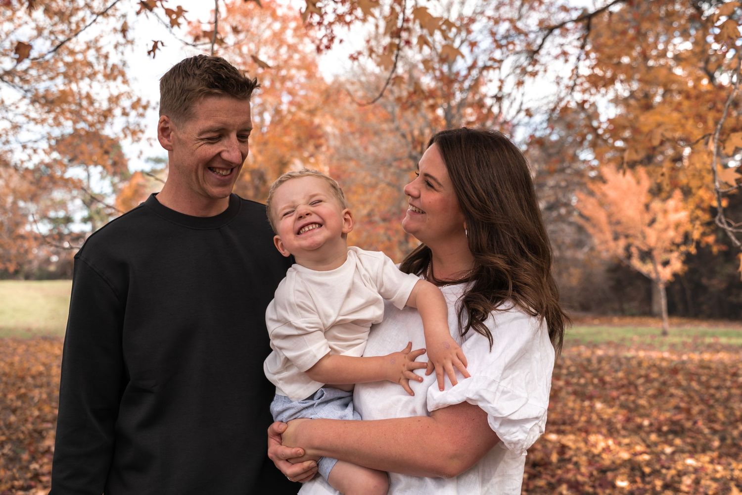 A happy family of three enjoying autumn in a park, parents smiling at their laughing toddler among golden fall foliage.
