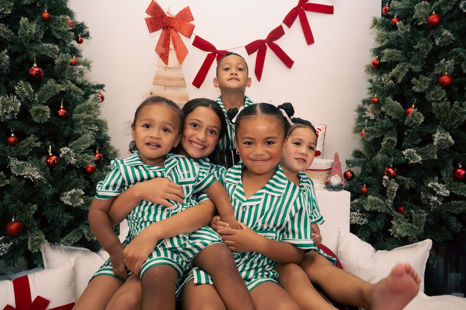 Five children in matching green striped pajamas pose together near Christmas trees with red bows and ornaments.
