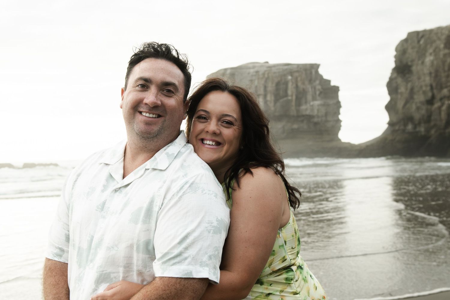 Smiling couple posing together on a rocky beach with dramatic sea cliffs in the background.