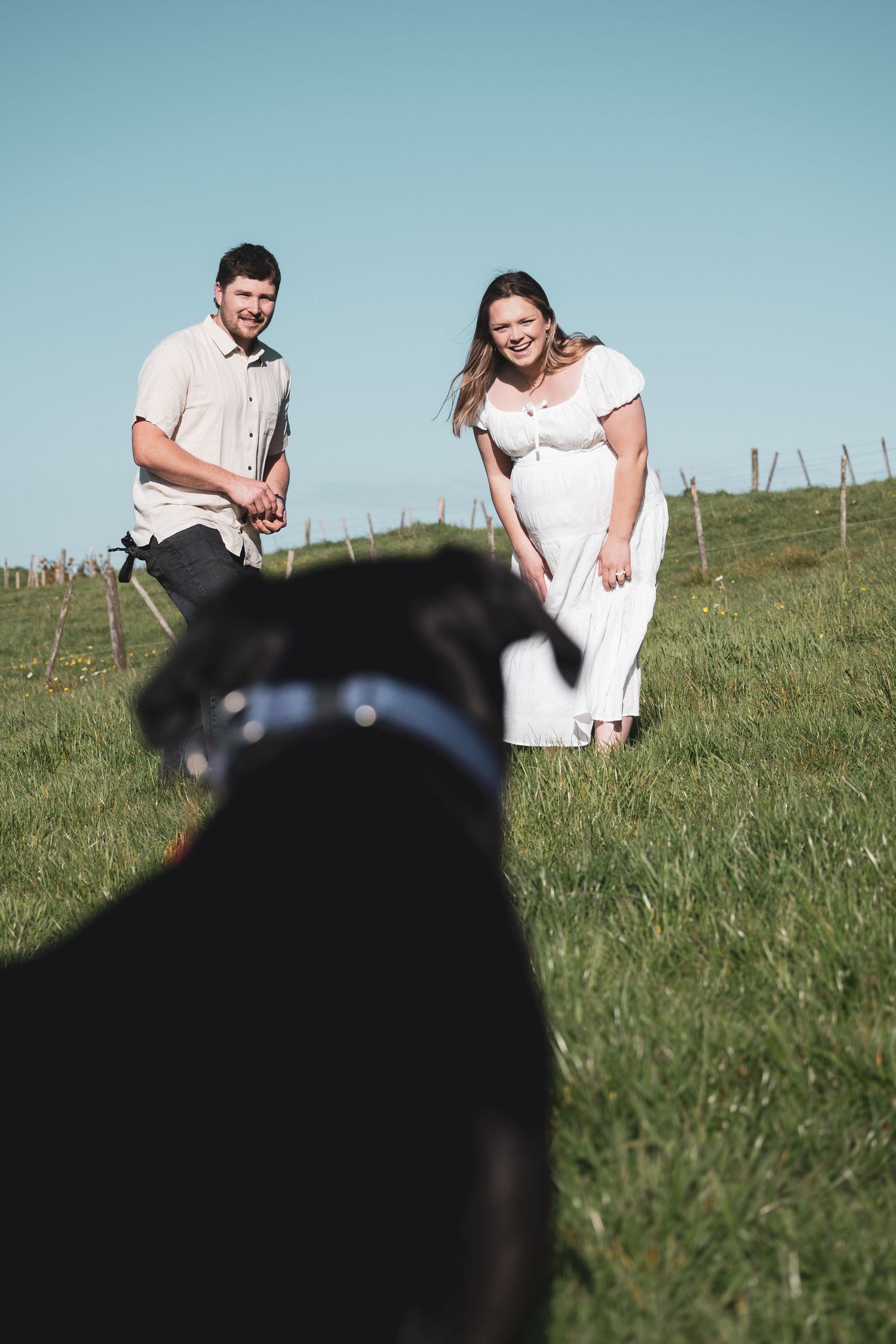 Pregnant woman in white dress and man smiling at black dog in green field under blue sky.