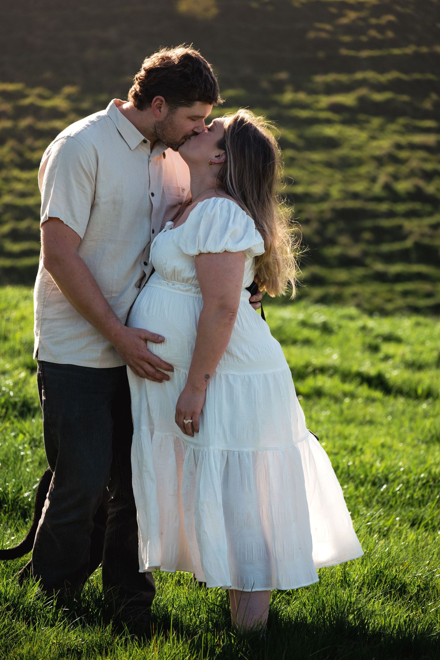 Couple sharing a tender kiss outdoors, pregnant woman in white dress with partner embracing her baby bump in sunlit green field.