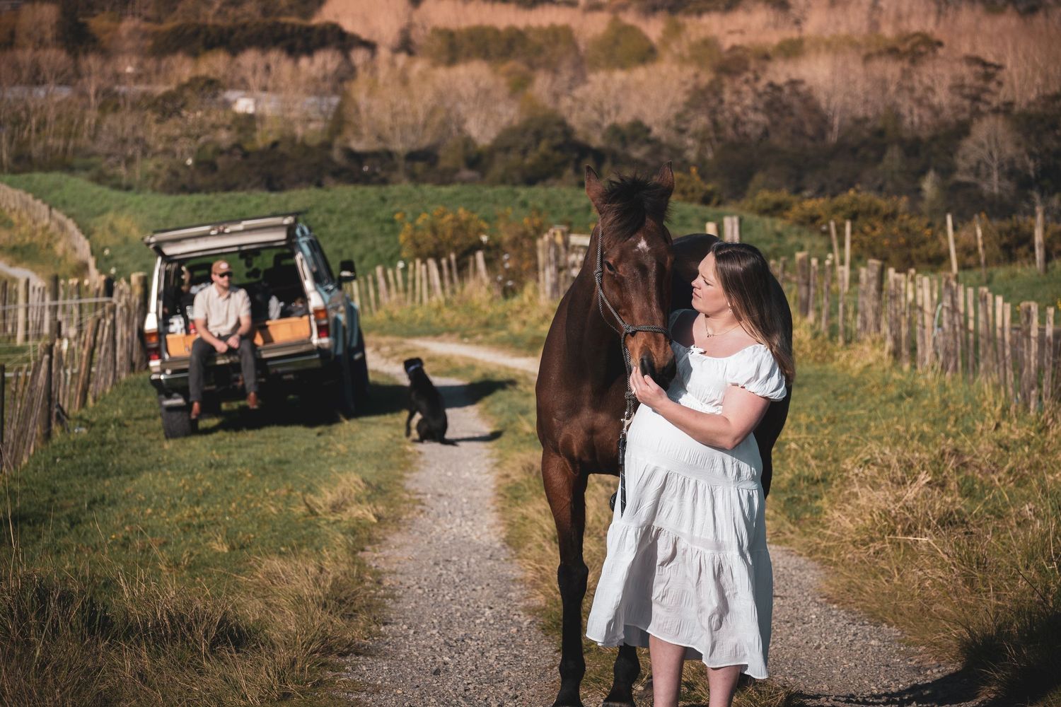 Woman in white dress holds brown horse on rural path beside parked SUV with man seated at back.