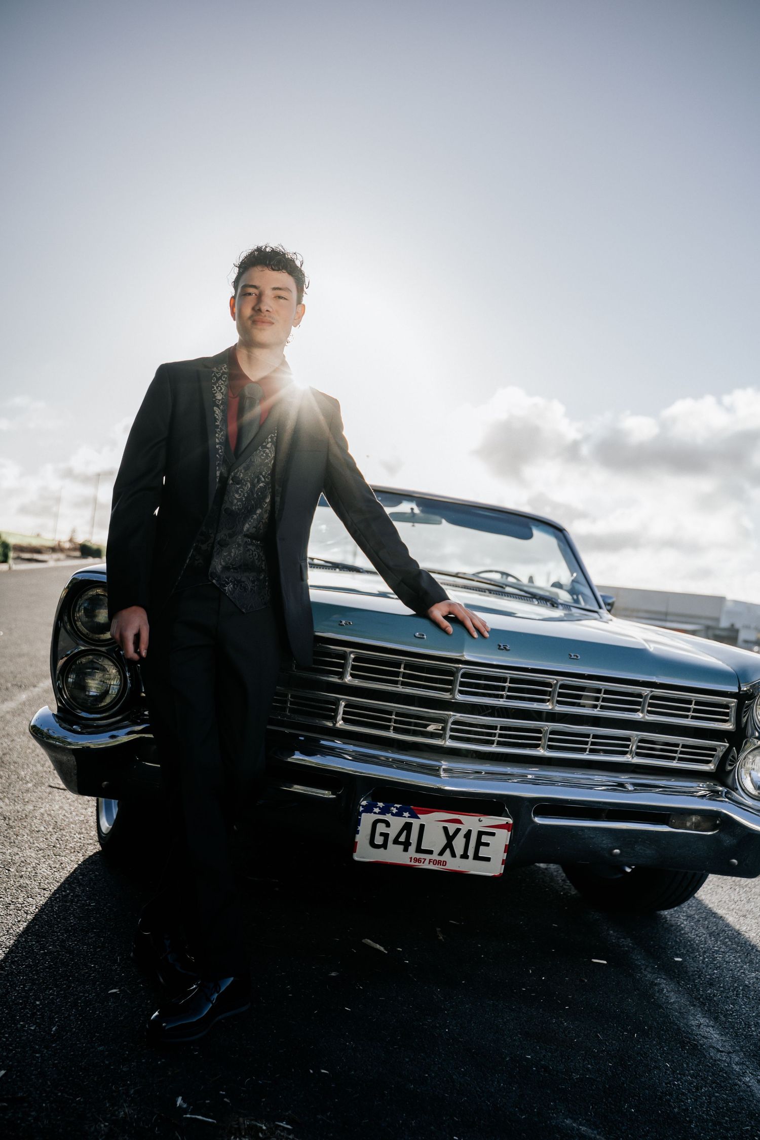 Young man in suit leaning on classic Ford Galaxie convertible with custom G4LXIE license plate in sunny parking lot.