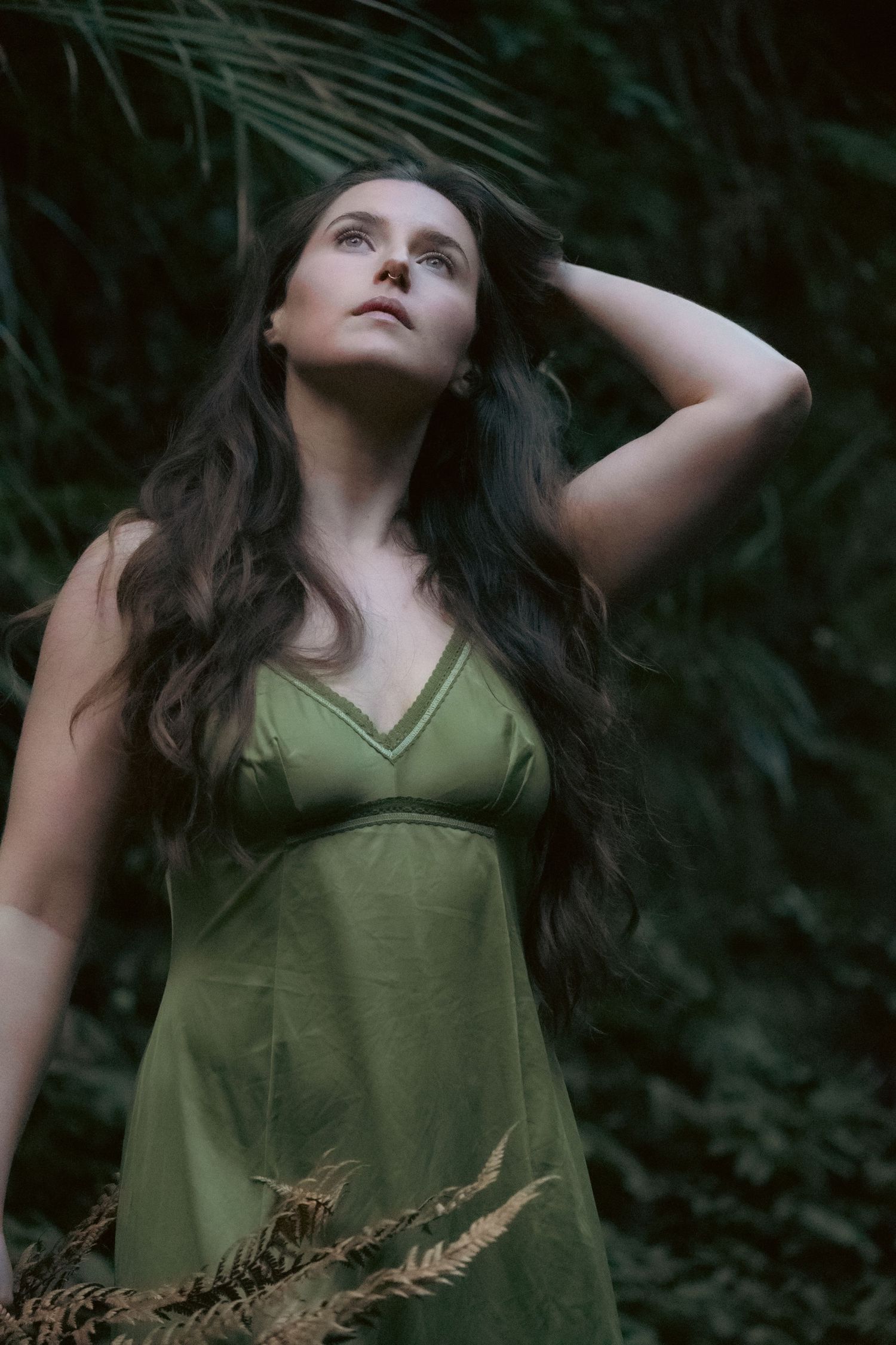 Woman in green dress gazes upward among lush tropical ferns and palm leaves in a serene forest setting.