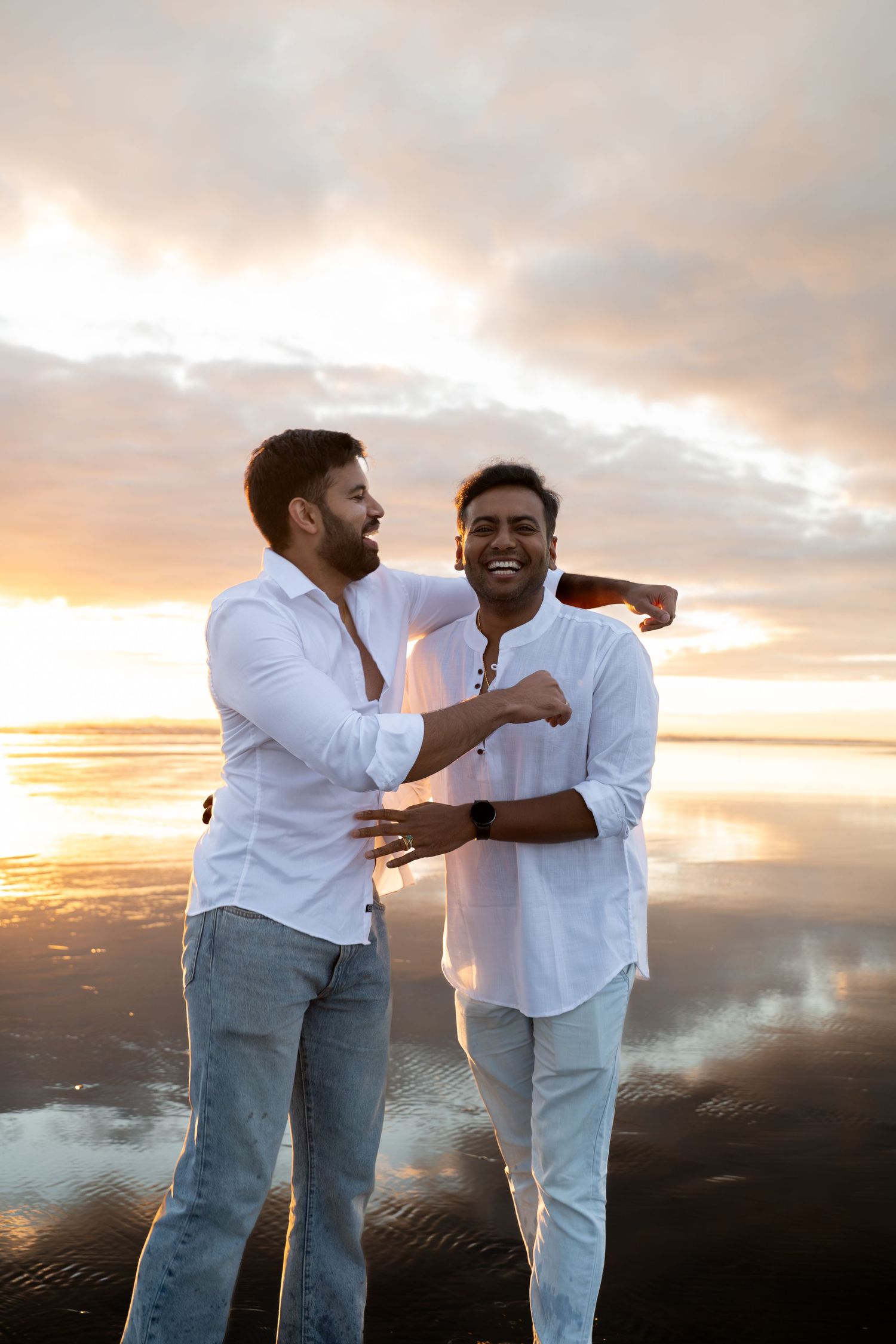 Two men in white shirts laughing together on a reflective beach at sunset.