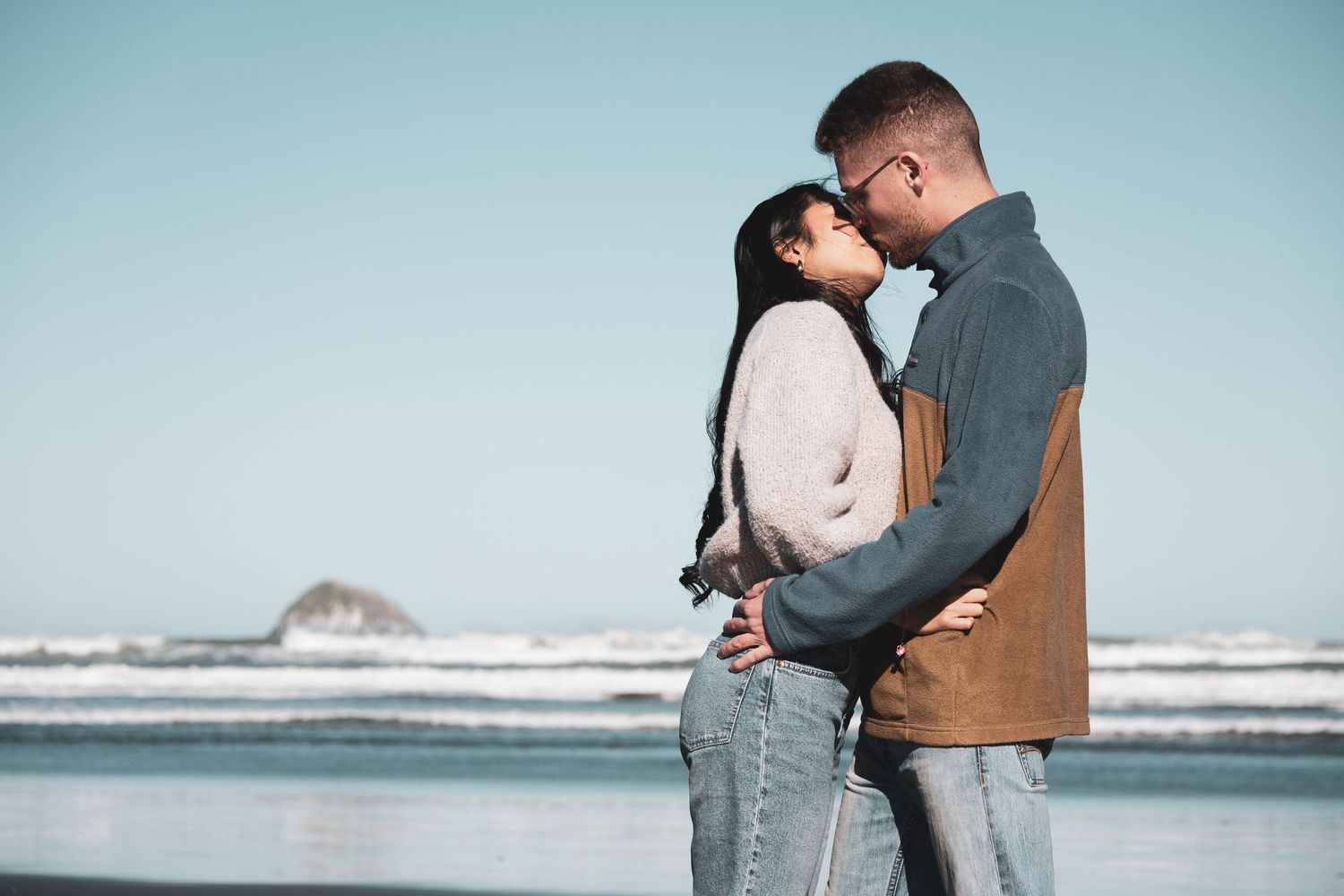 Romantic couple embracing on a beach with ocean waves and clear blue sky in the background.