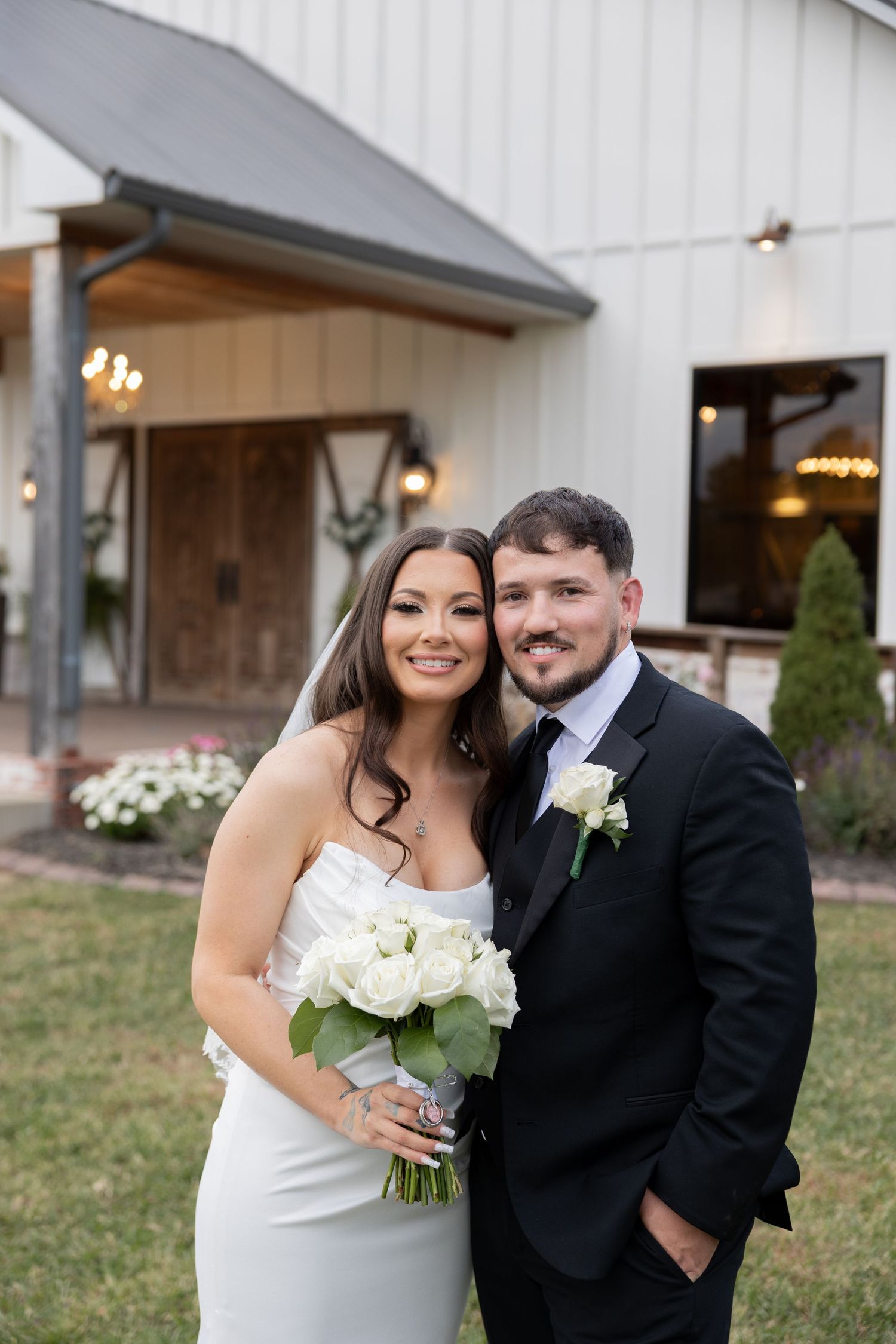 Bride and groom pose together outside a white barn wedding venue, bride holding white rose bouquet in elegant gown.