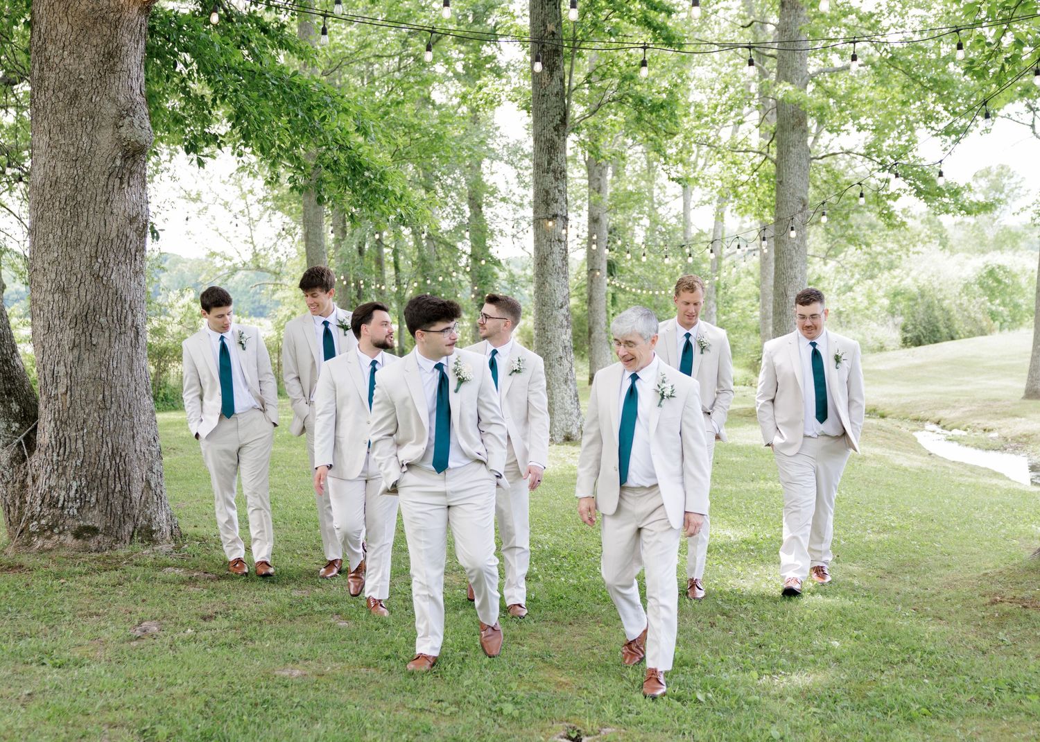 Groomsmen in tan suits with teal ties walking outdoors among tall trees on a sunny wedding day.