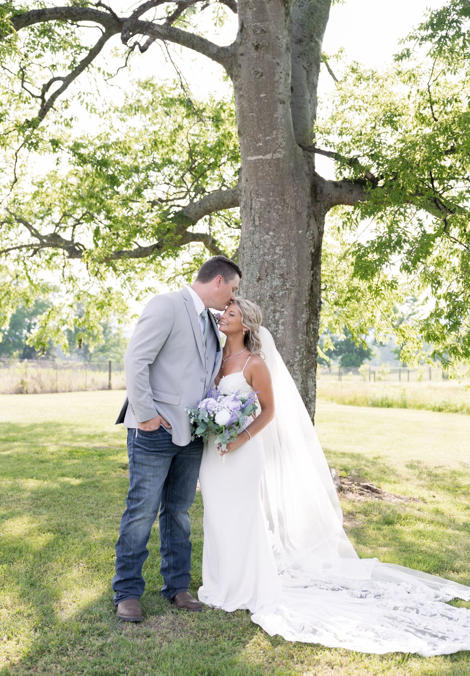 Bride and groom share a romantic kiss under a large oak tree, bride holding purple bouquet in white gown with veil.