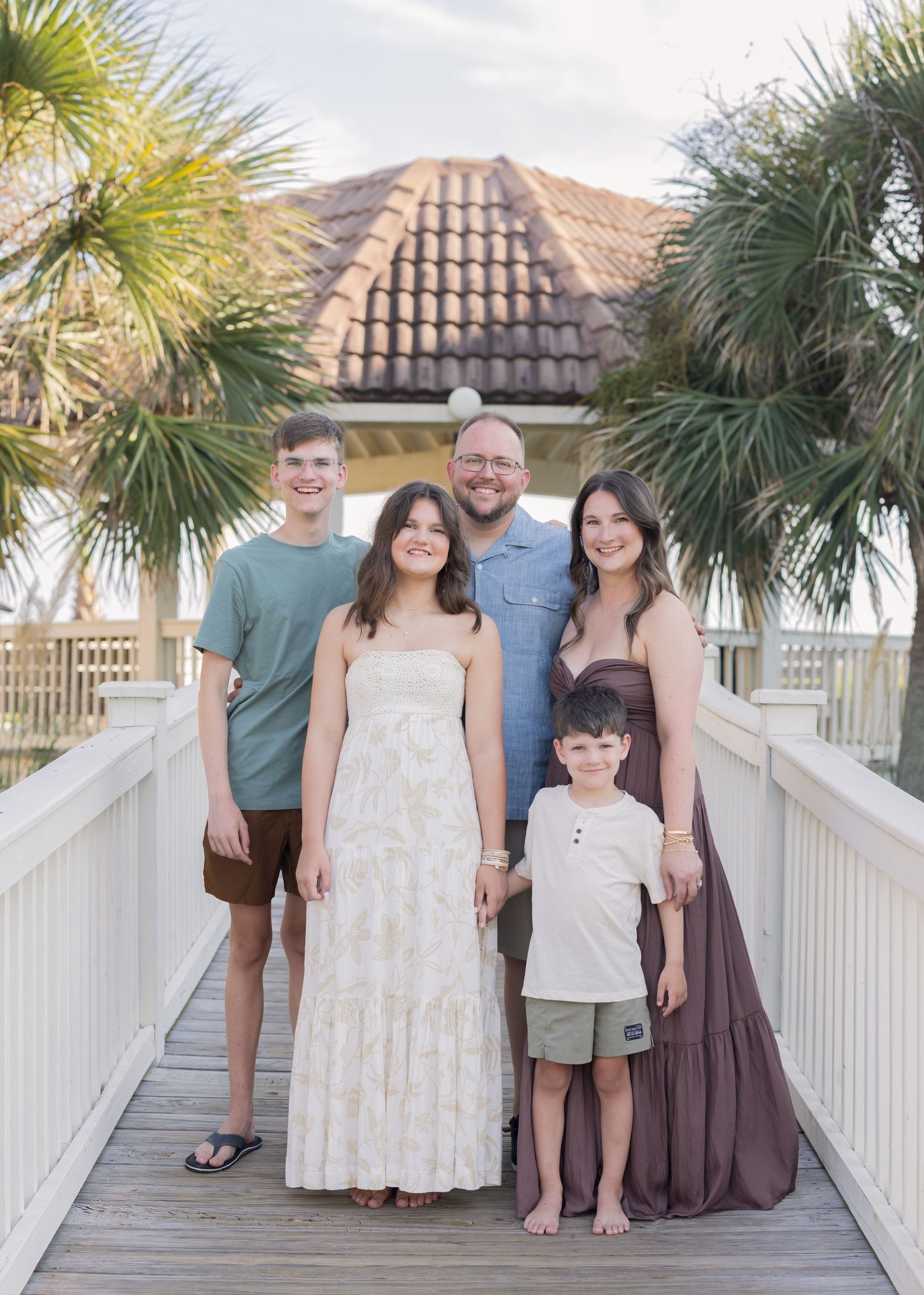 Family of five posing on a wooden boardwalk near a gazebo surrounded by palm trees at a beach resort.