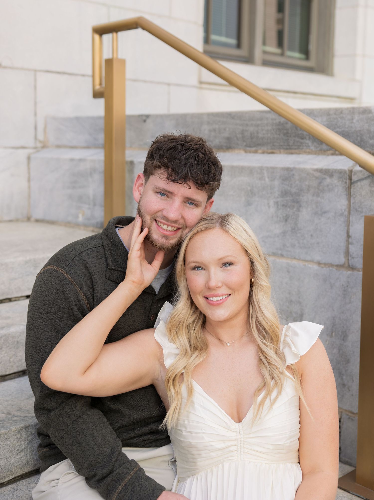 Smiling couple sitting on stone steps, woman in white dress, man in dark shirt, posing for engagement photo.