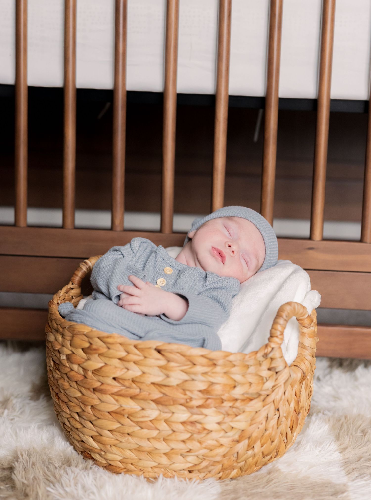 Peaceful newborn baby sleeping in a woven basket wearing blue outfit and hat beside a wooden crib.