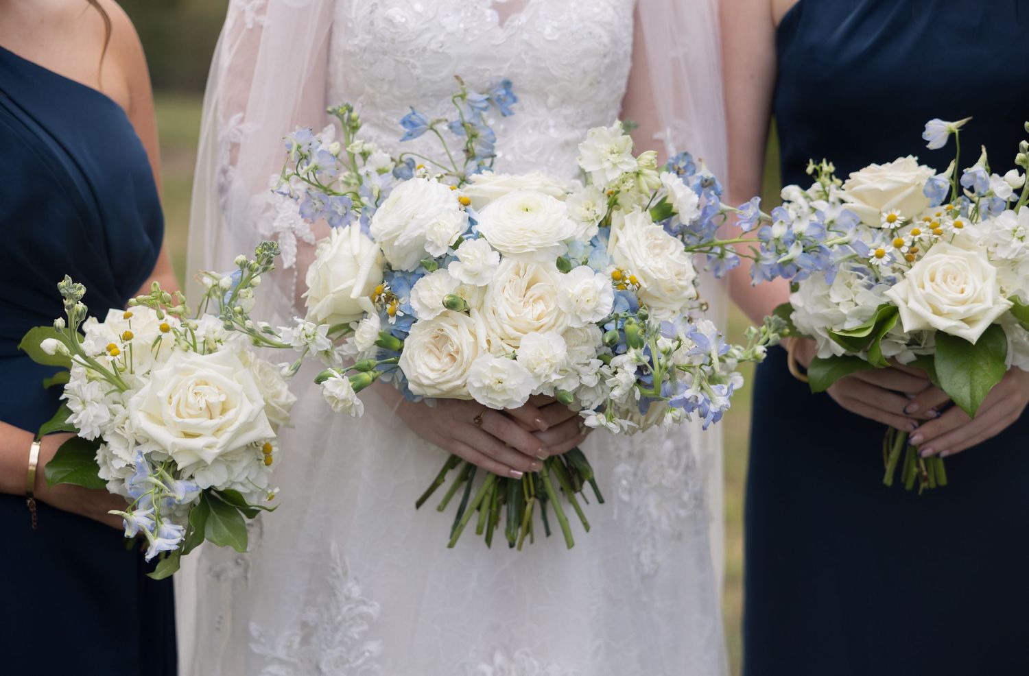 Bride and bridesmaids holding white rose and blue delphinium wedding bouquets, bridesmaids in navy dresses.
