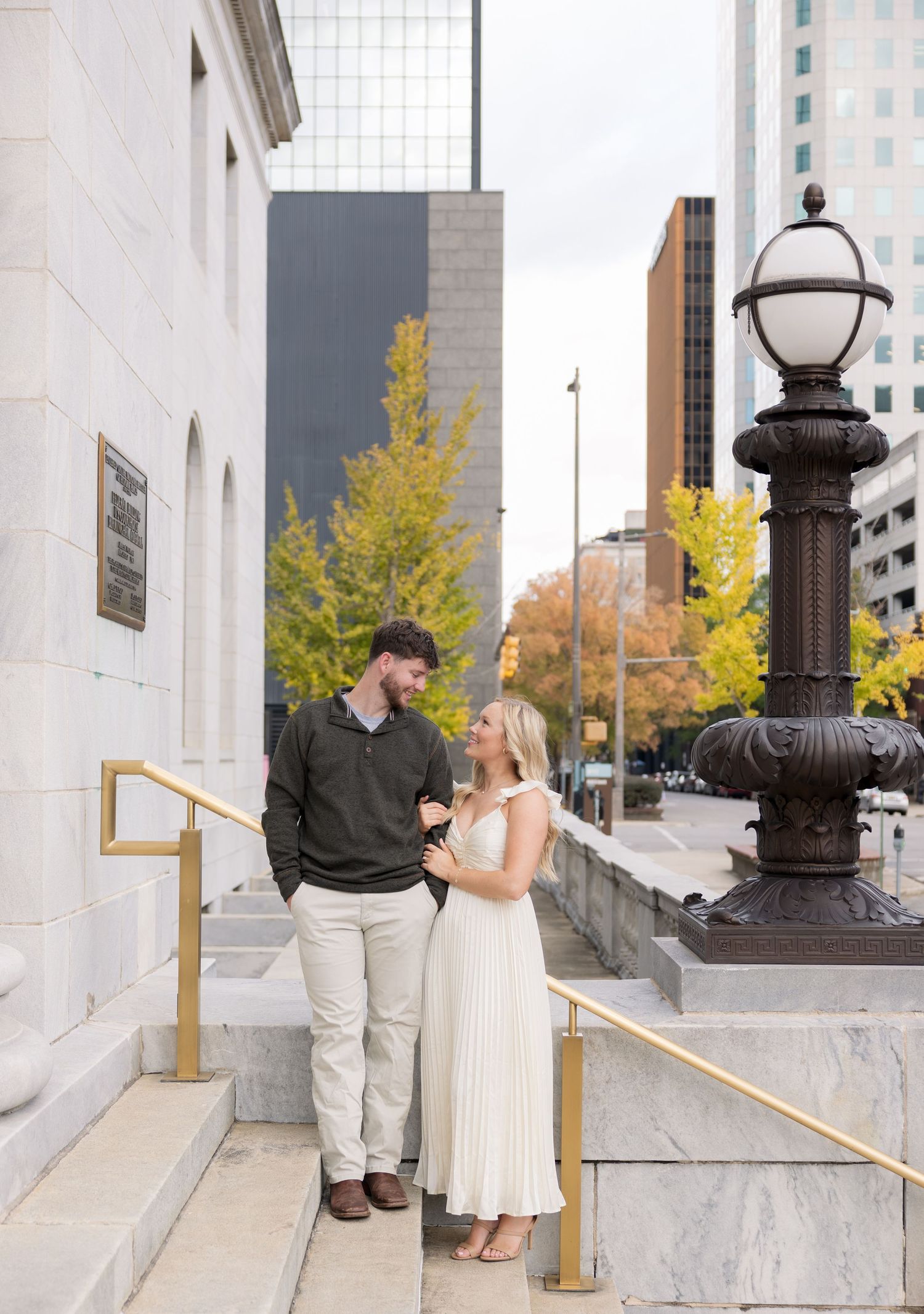 Couple sharing a romantic moment on stone steps outside a classic building with autumn trees and city skyline backdrop.
