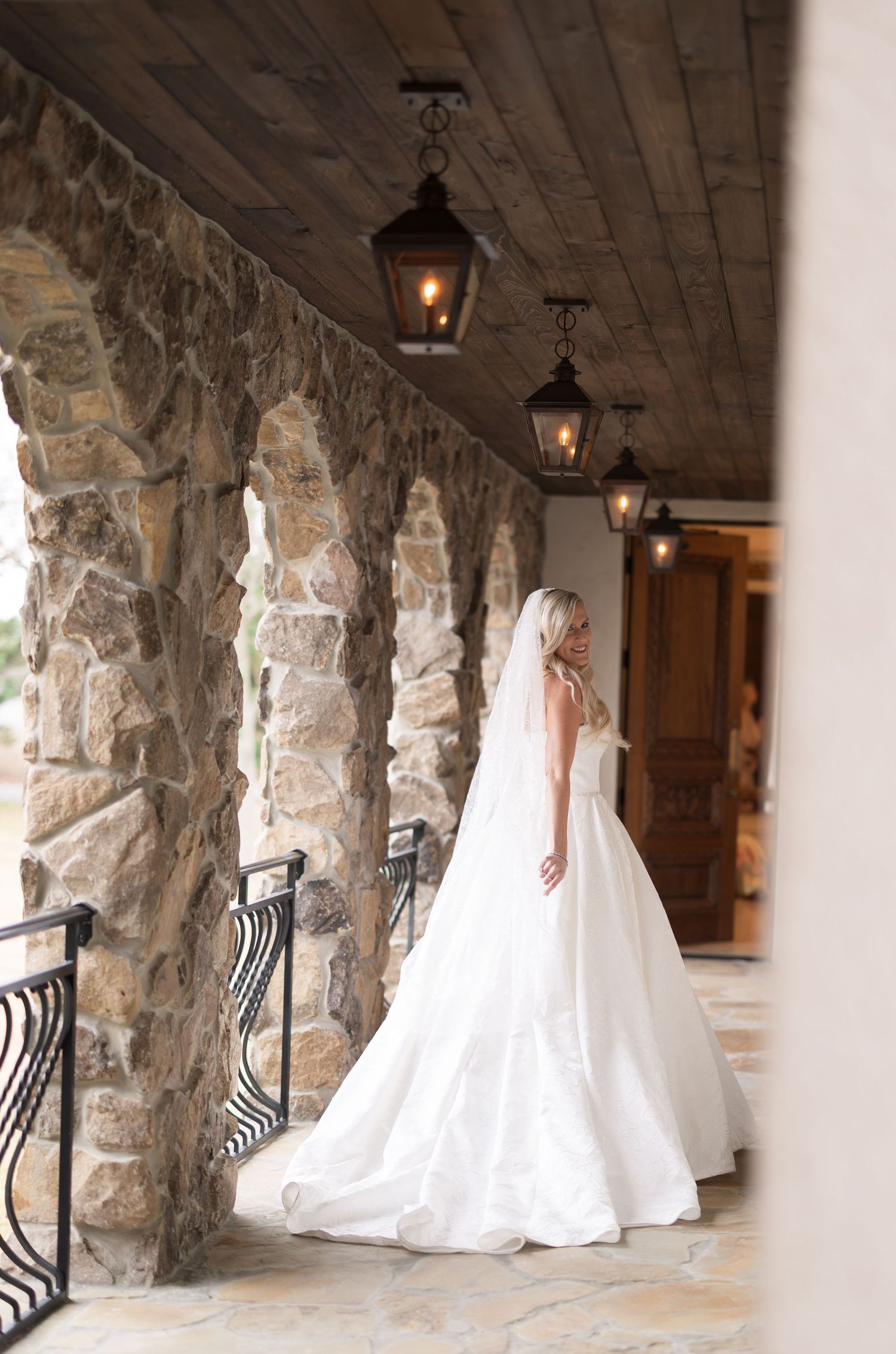 Bride in white ball gown and veil stands on stone archway corridor with vintage lanterns at elegant wedding venue.