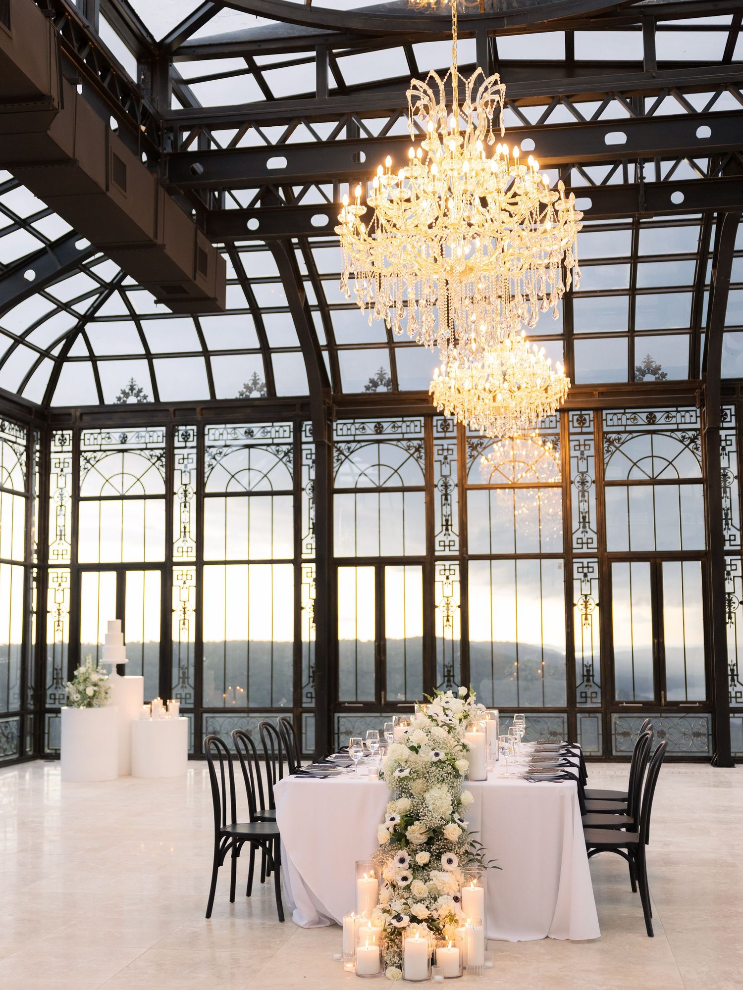 Elegant wedding reception in a glass conservatory with a grand crystal chandelier, white floral centerpiece, and candles at dusk.