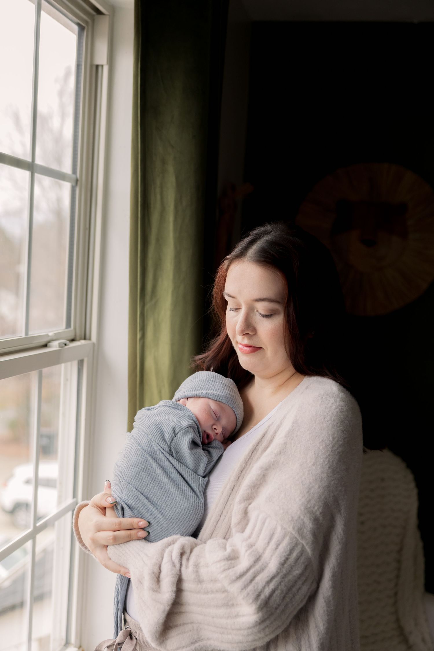 A young mother tenderly holds her swaddled newborn baby in a blue wrap near a sunlit window.