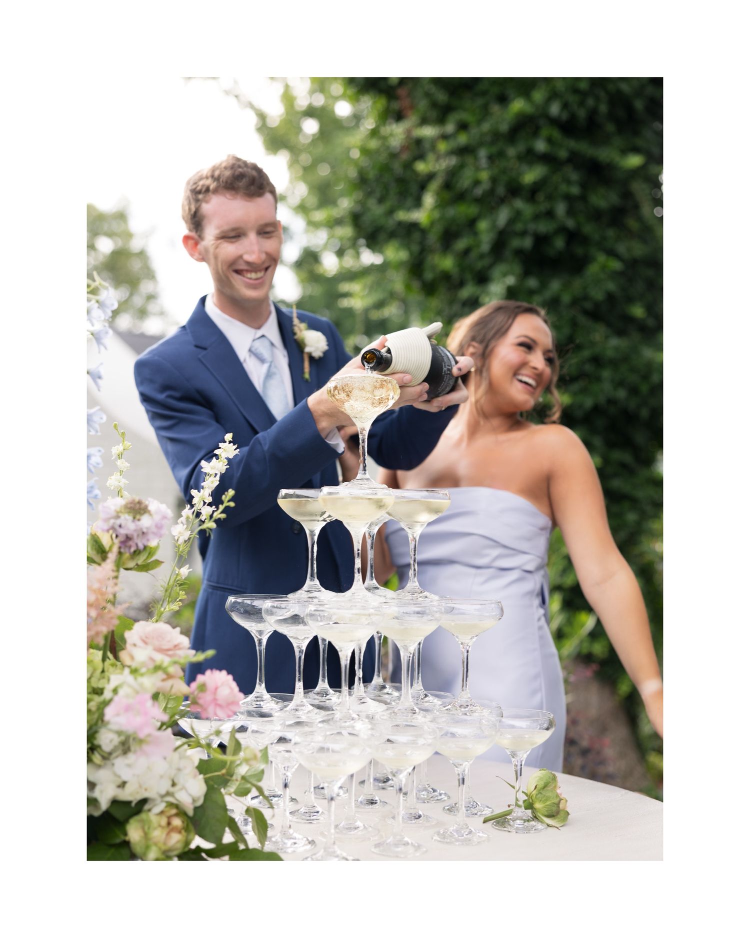 Bride and groom pouring champagne tower at outdoor wedding reception surrounded by floral arrangements.