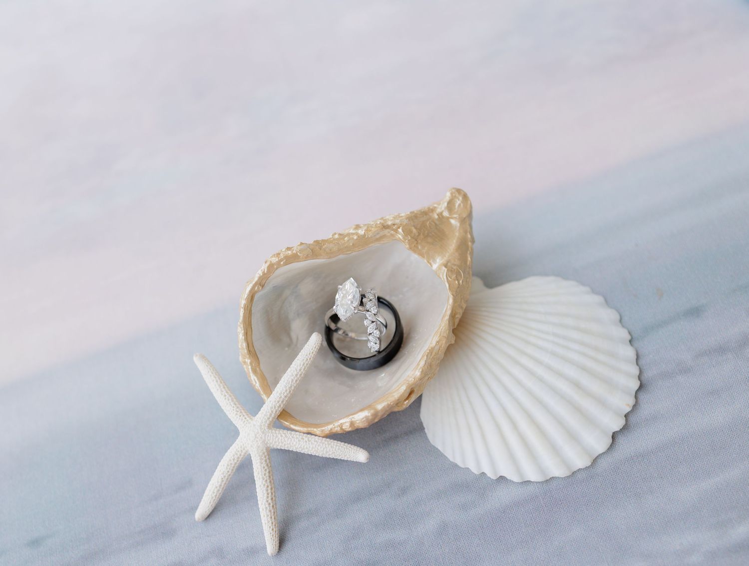 Wedding rings rest inside an open oyster shell beside a white starfish and scallop shell on a soft pastel beach-themed backdrop.