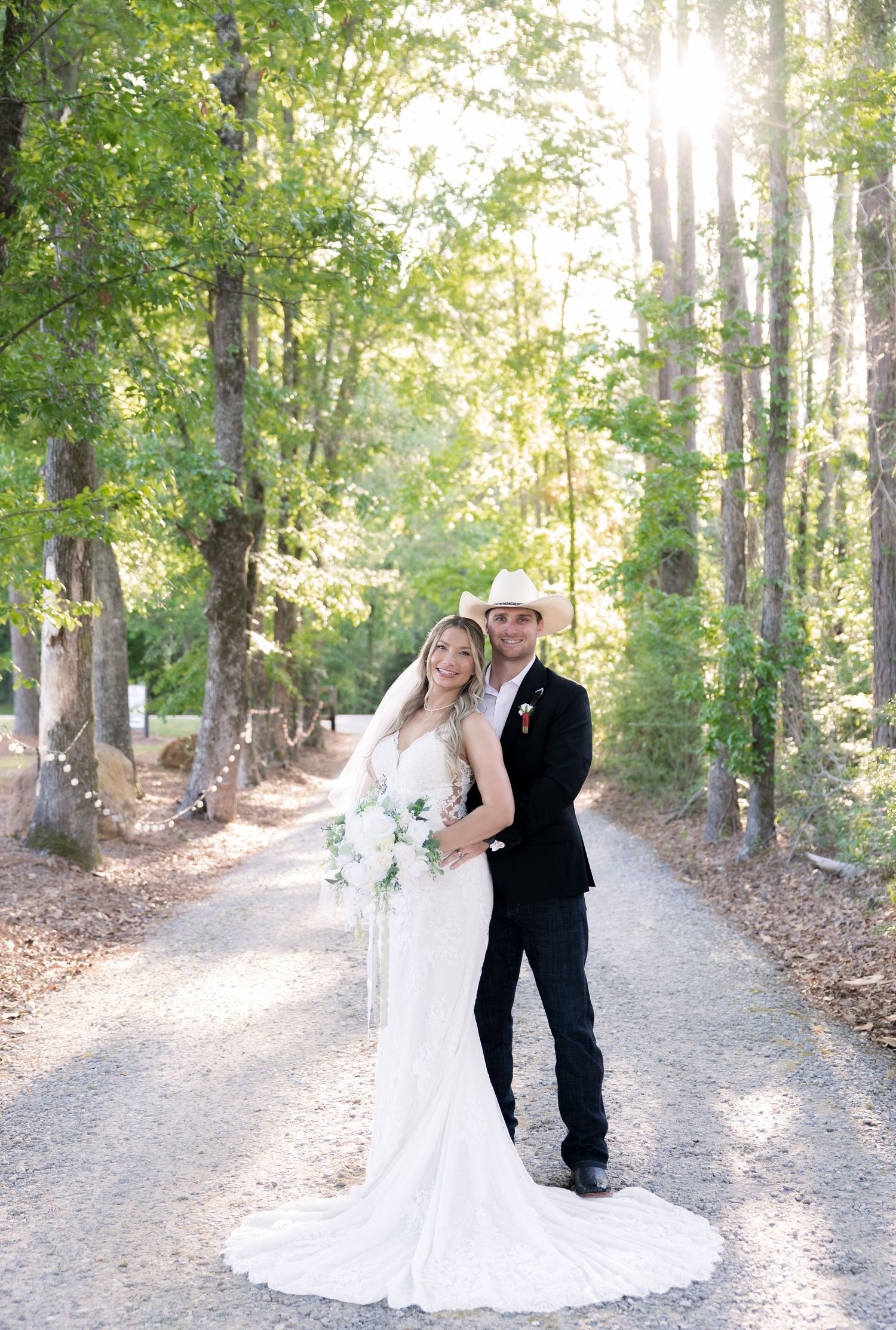 Bride and groom pose on a sunlit gravel path surrounded by lush trees, she in a white gown, he in a black suit with cowboy hat.