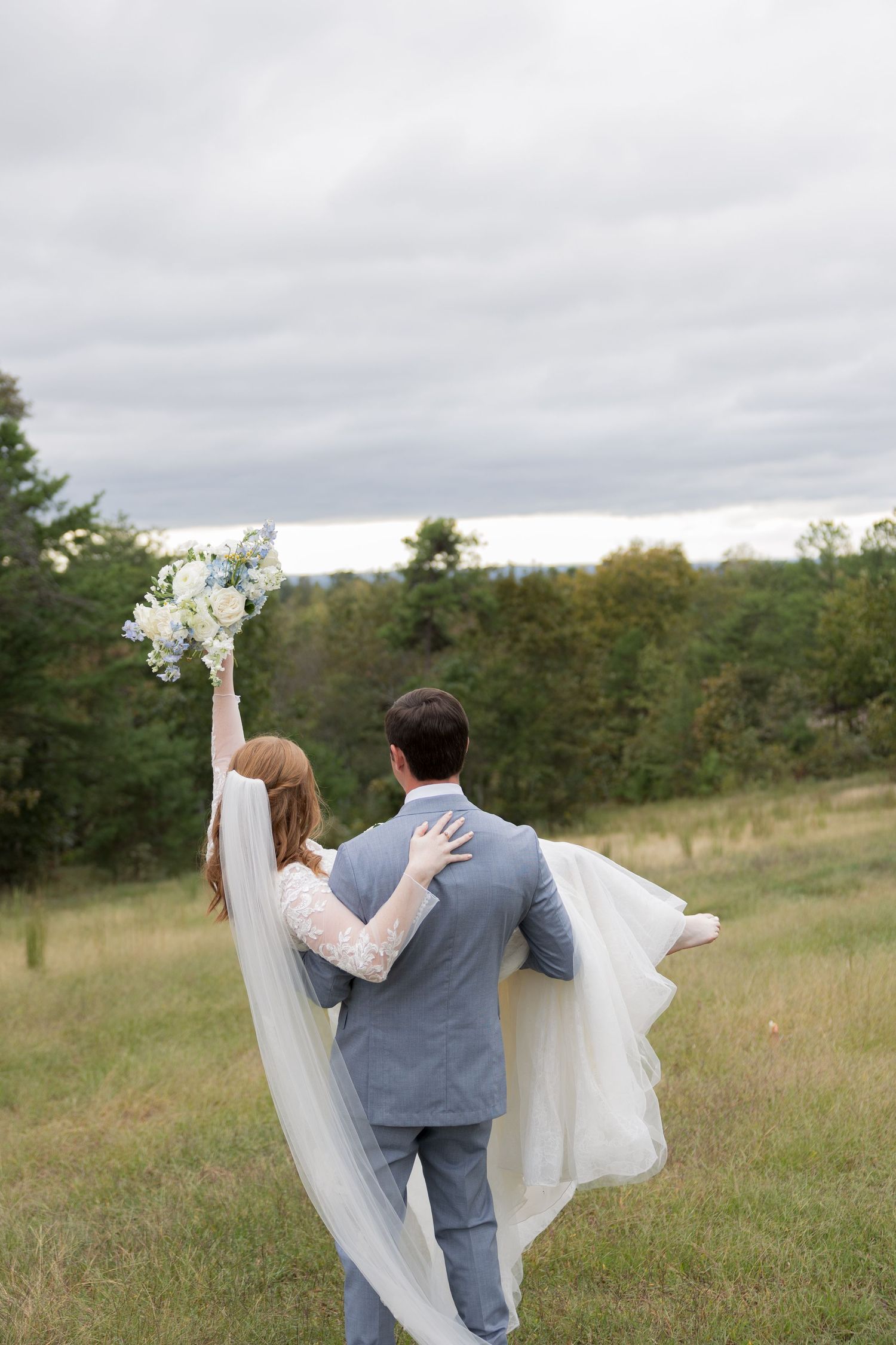 Bride and groom walking through a green meadow, bride raising bouquet of white and blue flowers, wearing a flowing veil.