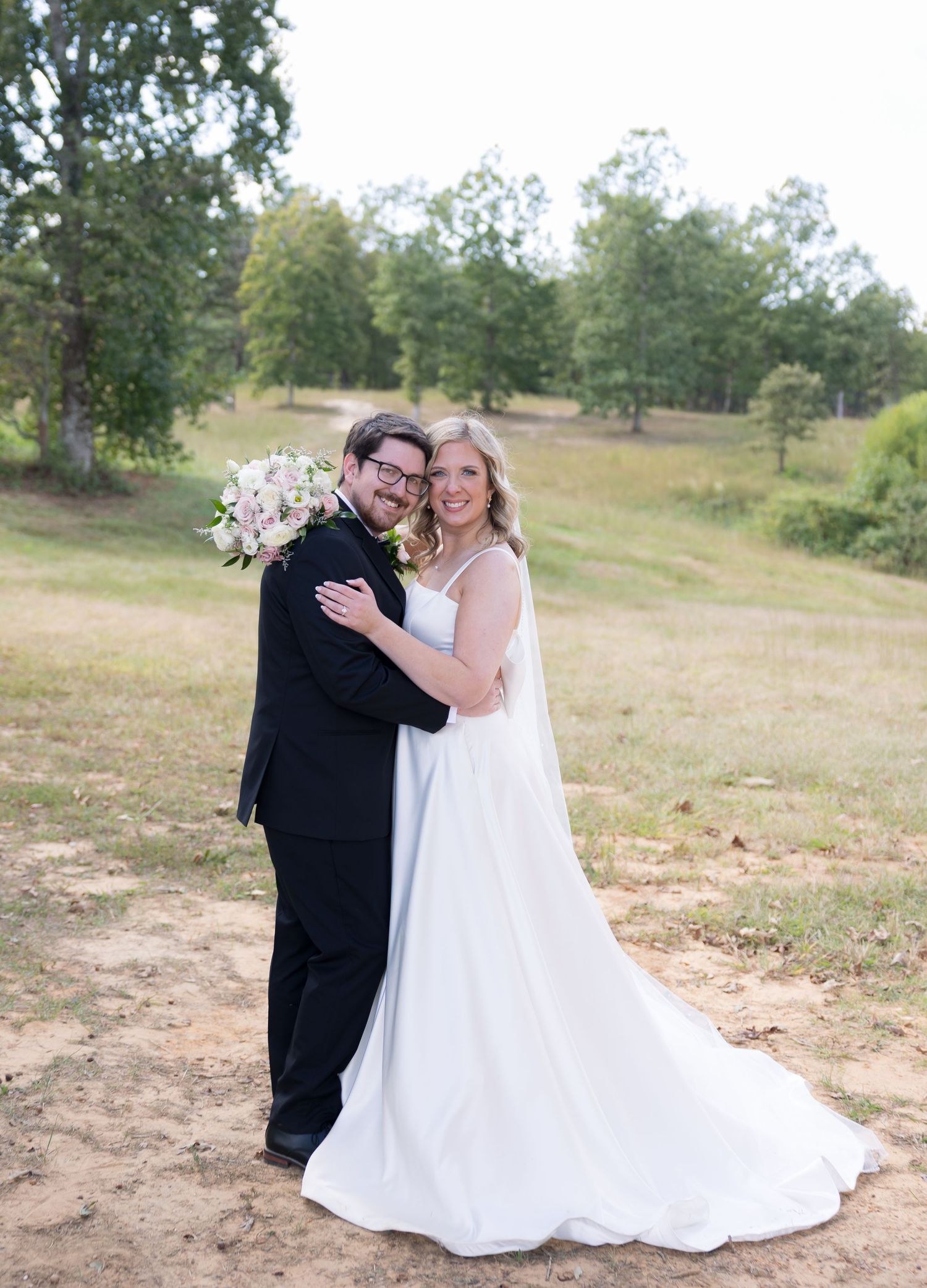 A bride in a white gown and groom in a black tuxedo pose together outdoors on their wedding day.