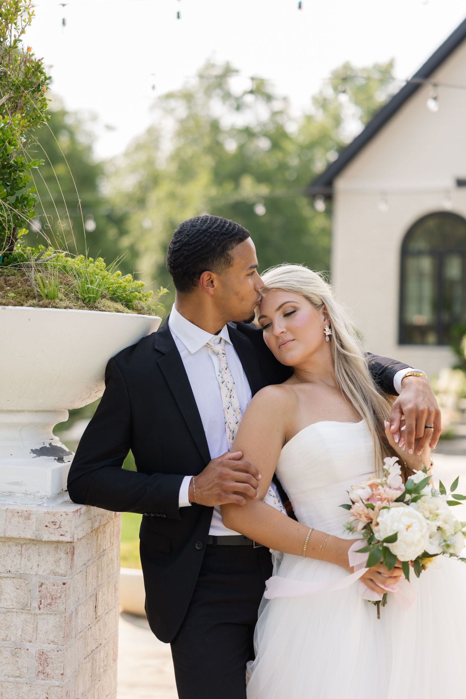 Groom in black suit kisses bride's temple as she leans on him holding a blush bouquet outdoors.