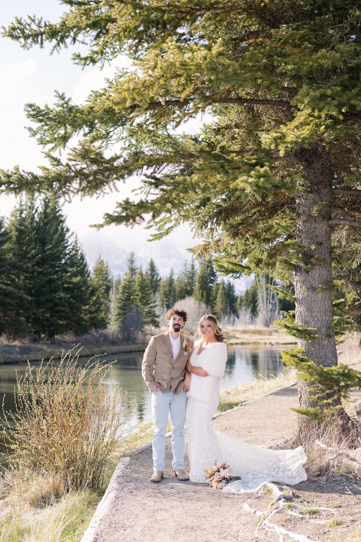 A bride and groom pose by a serene mountain lake surrounded by tall pine trees on their wedding day.