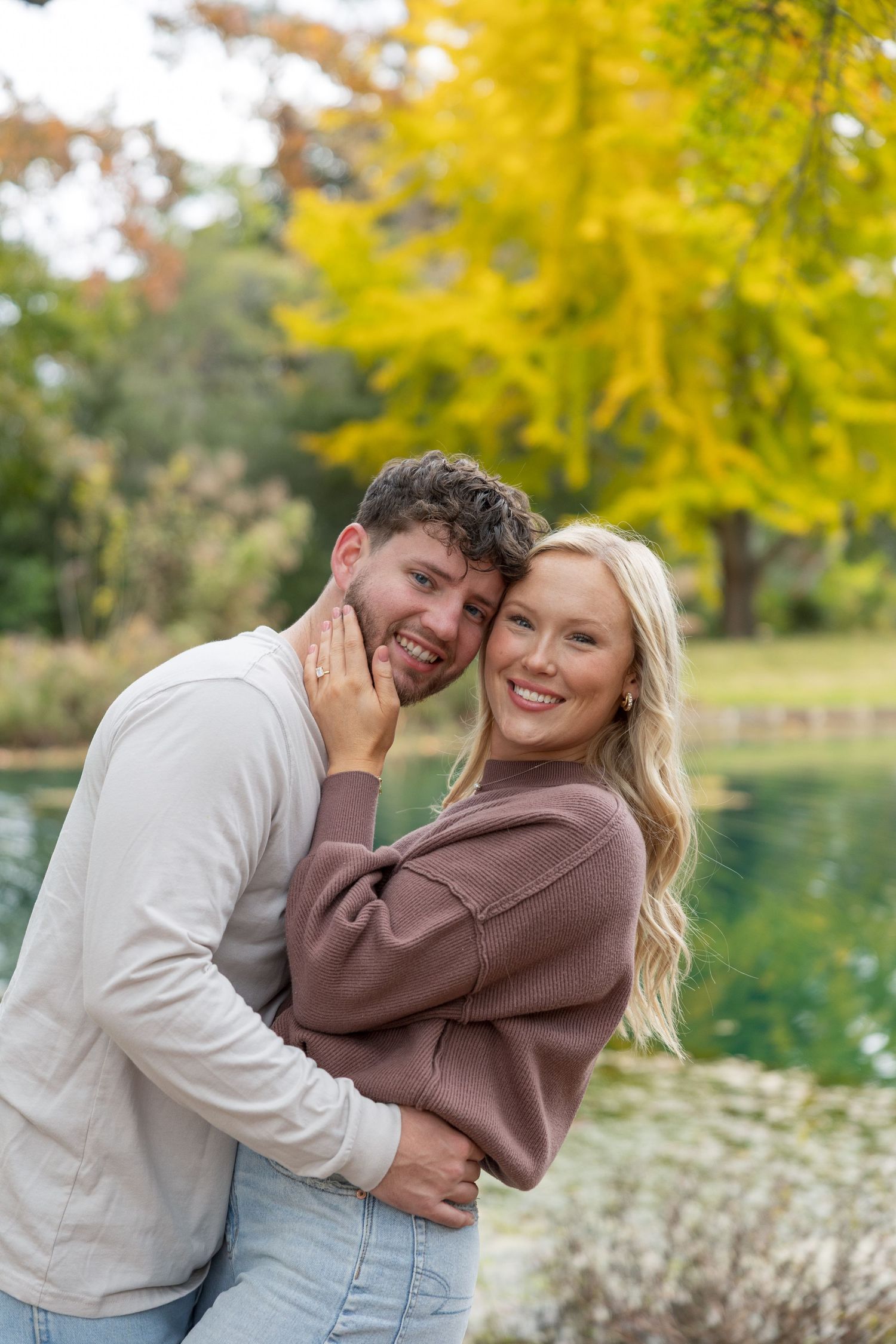 Happy couple embracing outdoors near a pond with vibrant yellow autumn trees in the background.