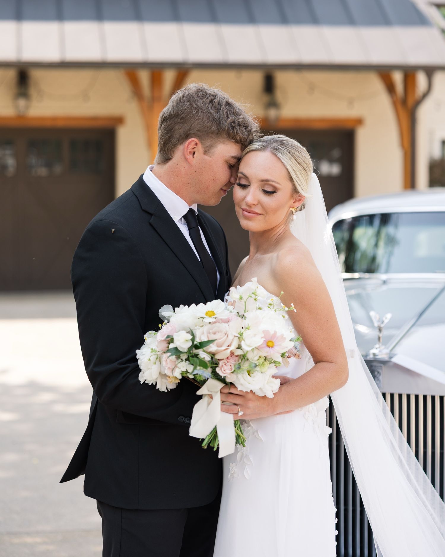 Bride and groom share a tender moment, she holds a white and pink bouquet, wearing a strapless gown with veil outdoors.