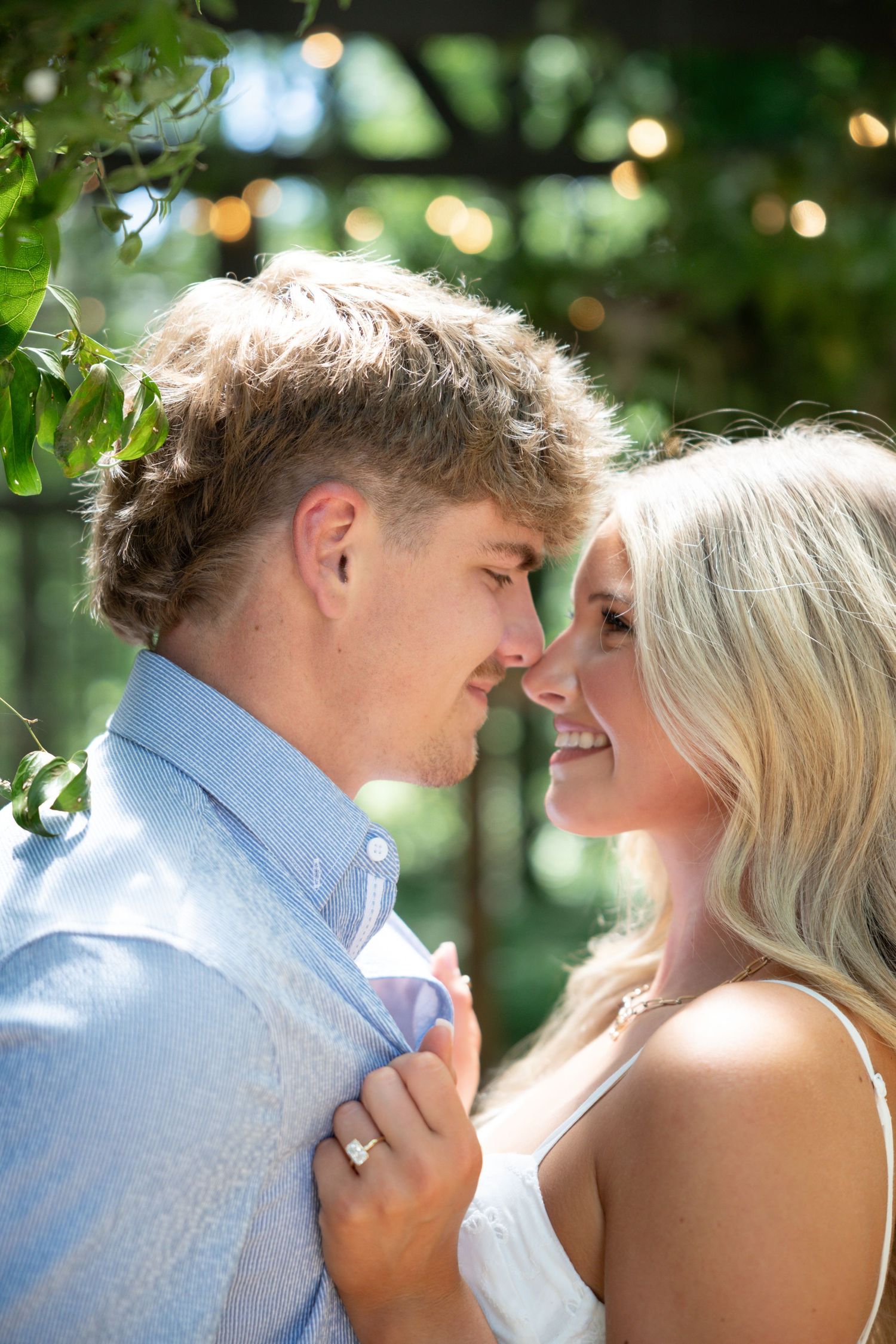 Couple sharing a romantic moment outdoors, woman holding man's shirt showing engagement ring, surrounded by lush greenery.