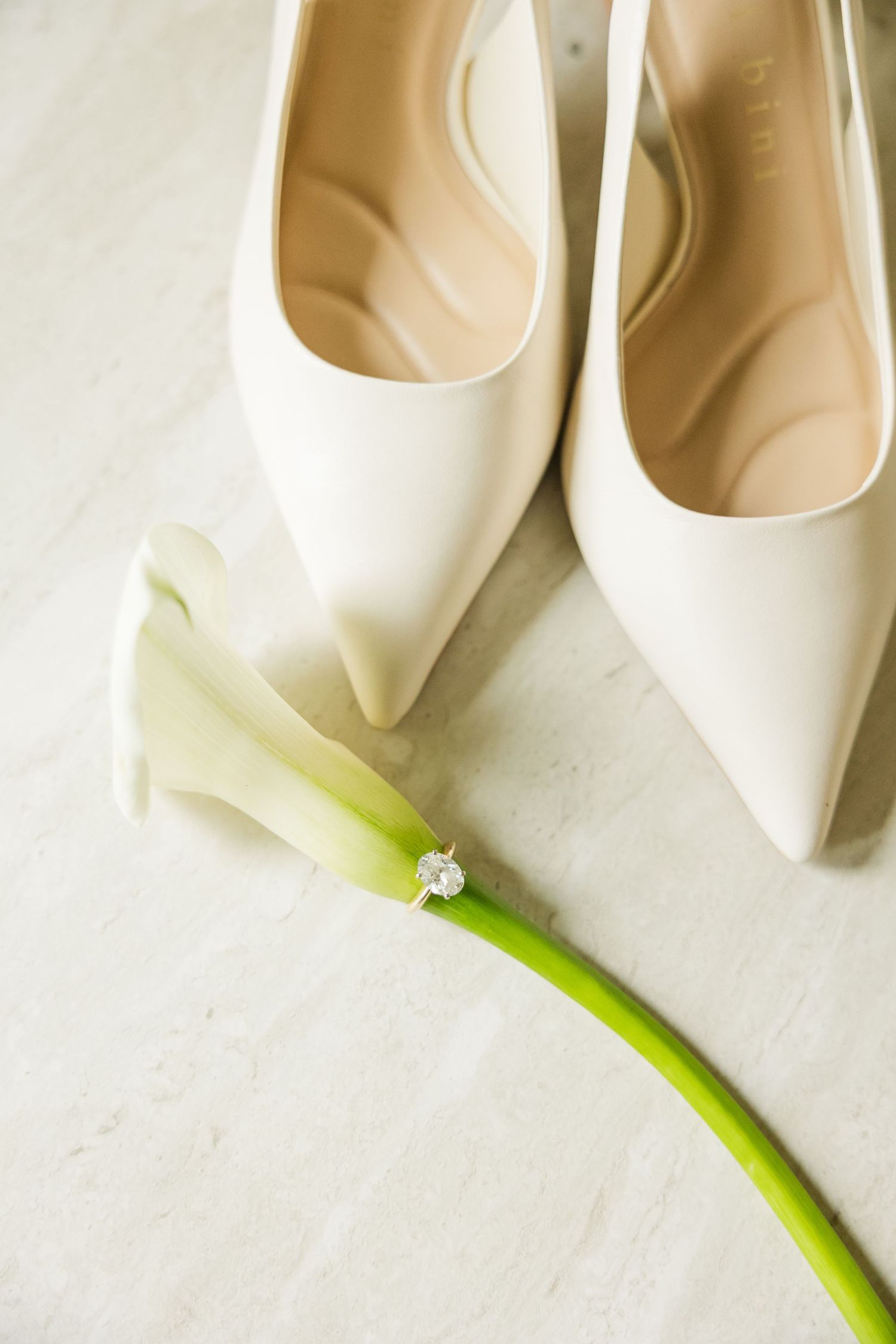 White bridal heels and diamond engagement ring resting on a calla lily on a light marble surface.