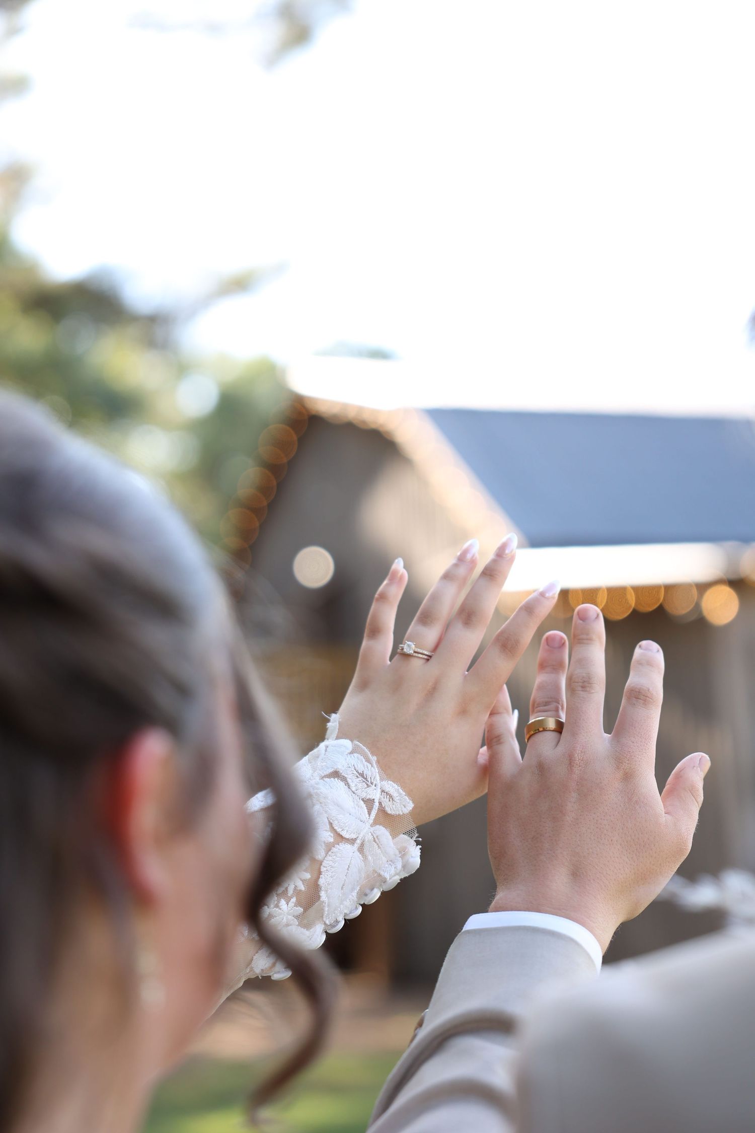 Newlyweds displaying wedding rings on their fingers outdoors at a wedding venue with warm bokeh lights.