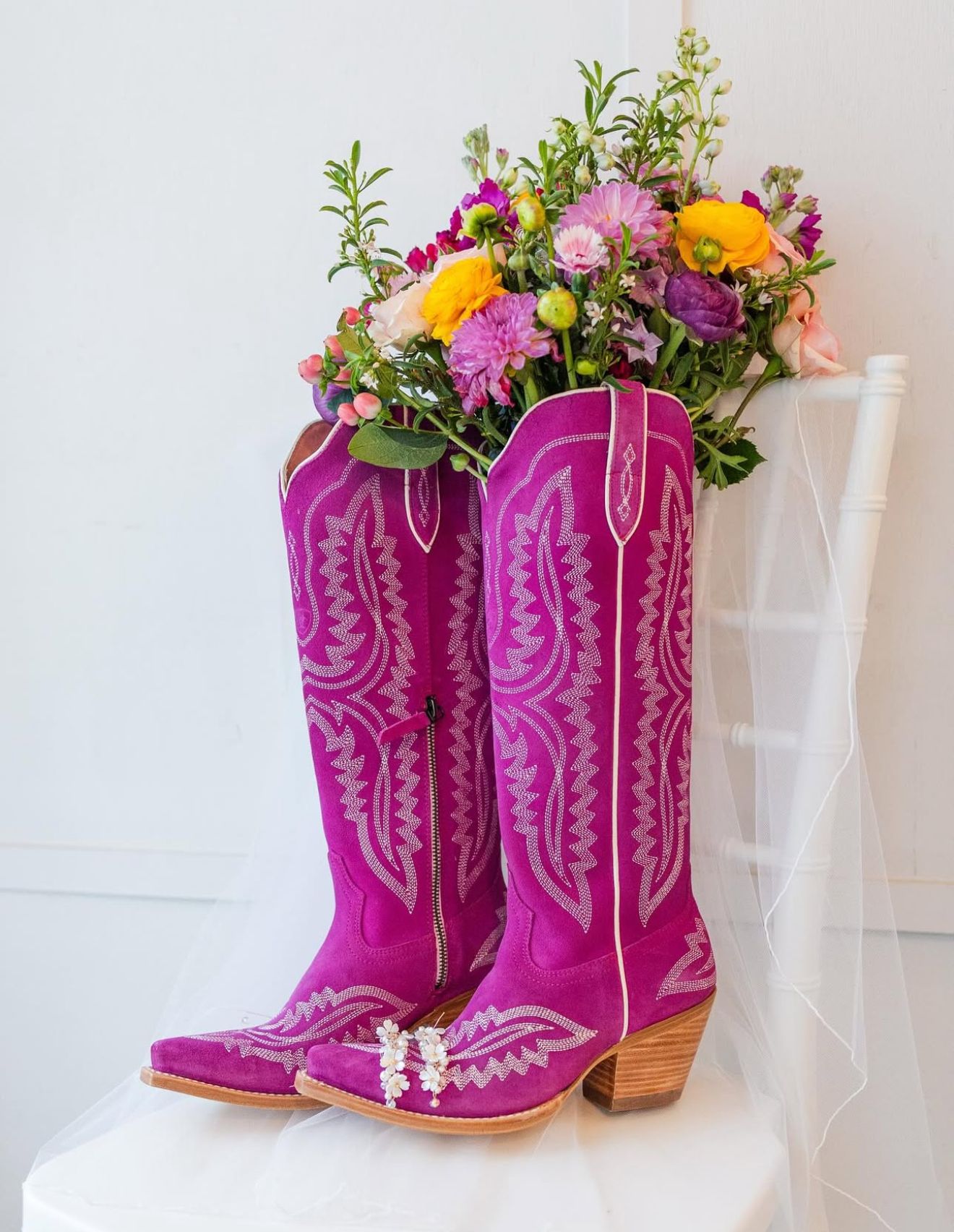 Vibrant magenta cowboy boots filled with colorful wildflowers, displayed on a white chair with a bridal veil.