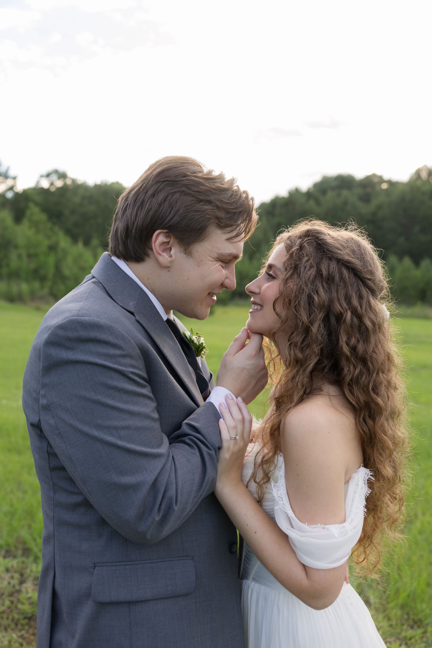 A groom in a gray suit tenderly holds a smiling bride in a white dress outdoors in a lush green field.