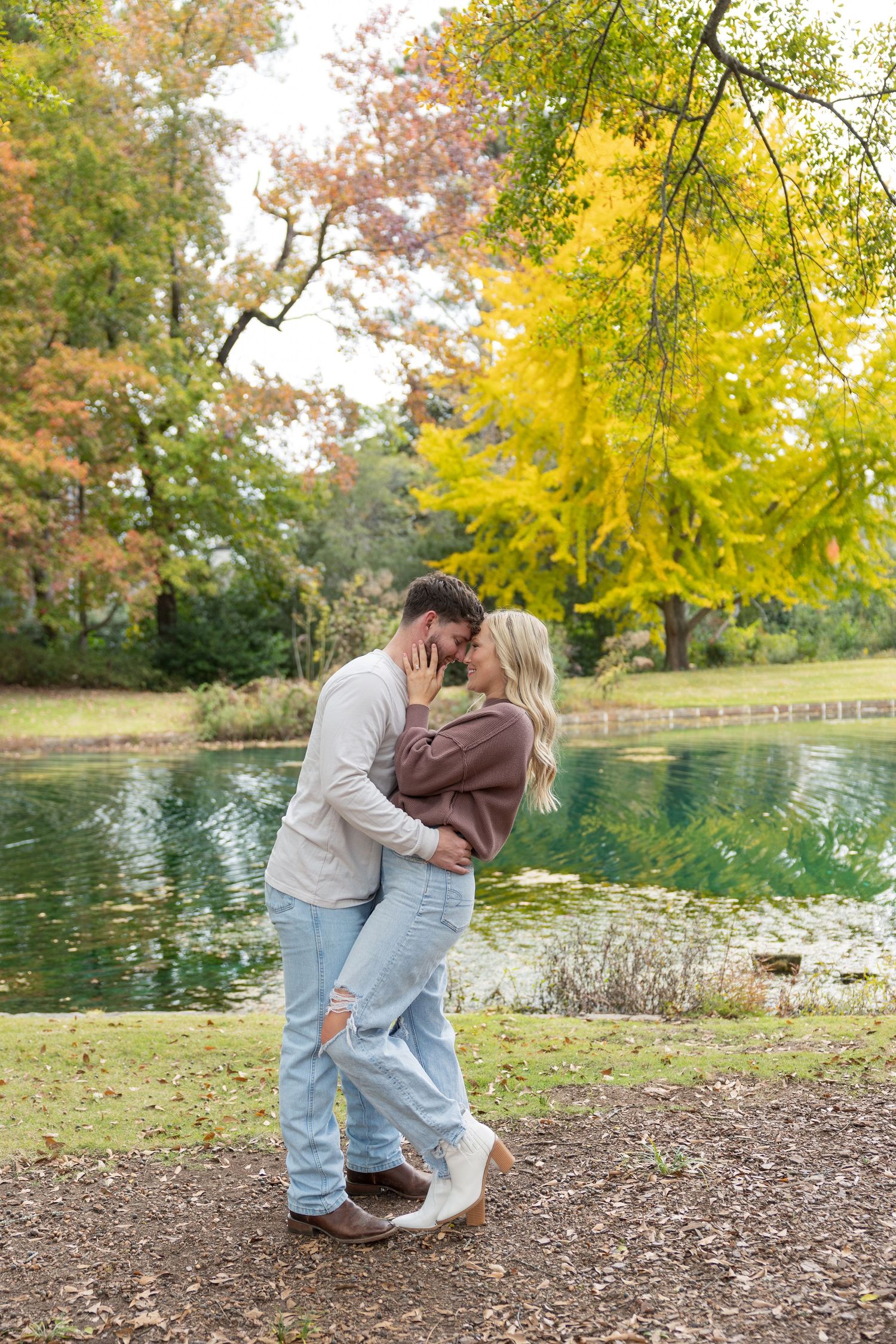 Couple sharing a romantic kiss by a serene pond surrounded by vibrant autumn foliage in a park.