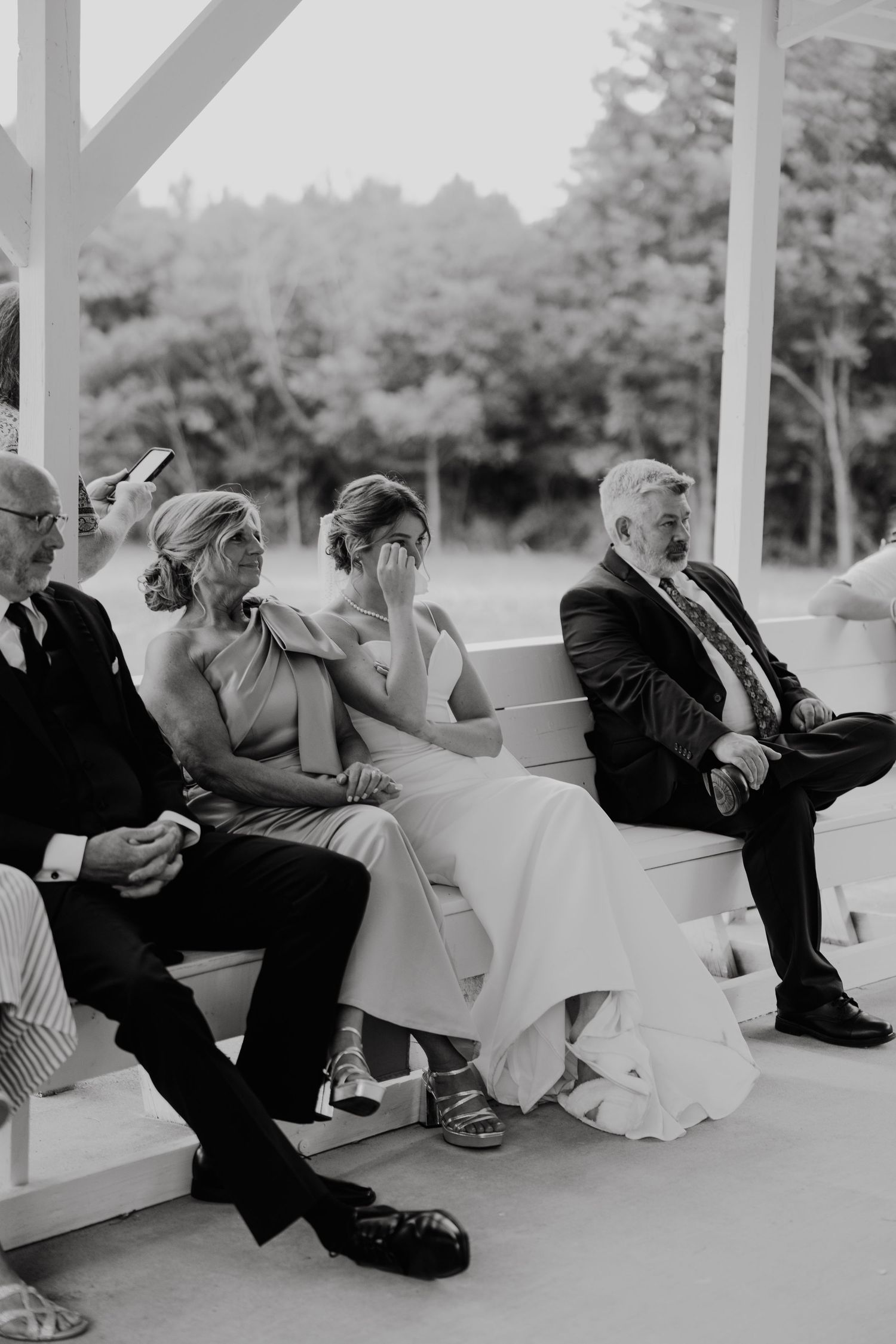 Black and white photo of emotional wedding guests seated on benches, with a woman wiping tears during an outdoor ceremony.
