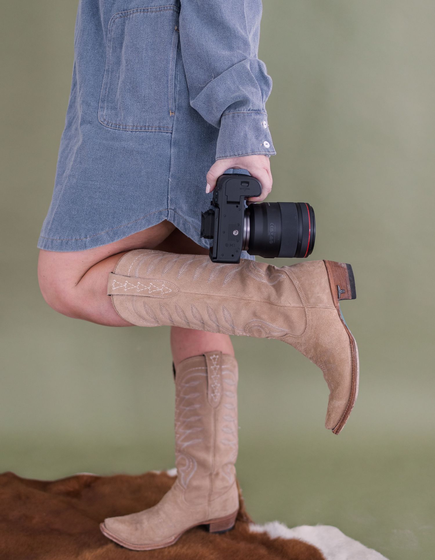 Photographer in denim dress and tan cowboy boots holds a DSLR camera against a sage green background.