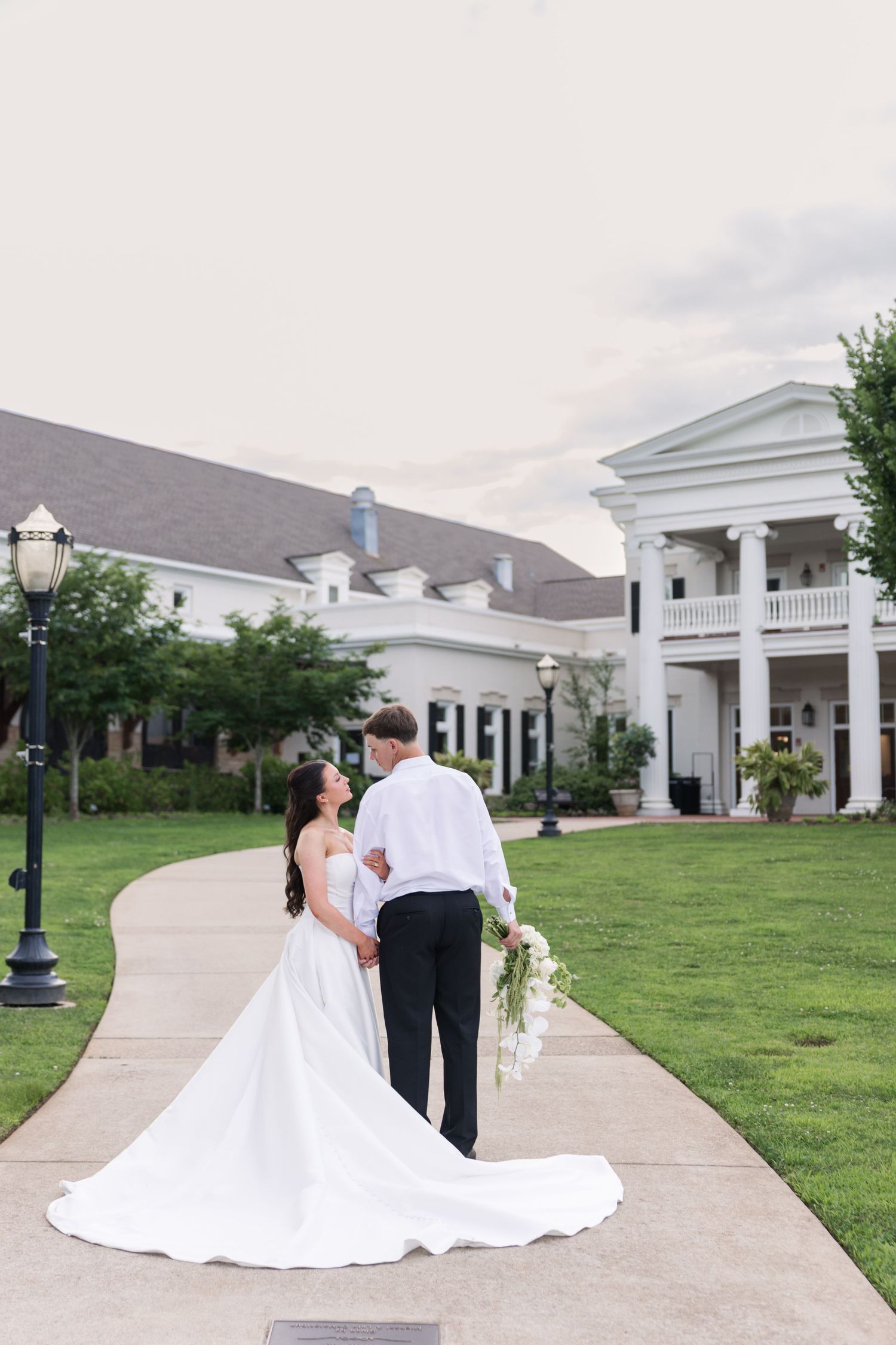 A bride and groom walk hand-in-hand on a pathway toward a grand white colonial venue surrounded by lush greenery.