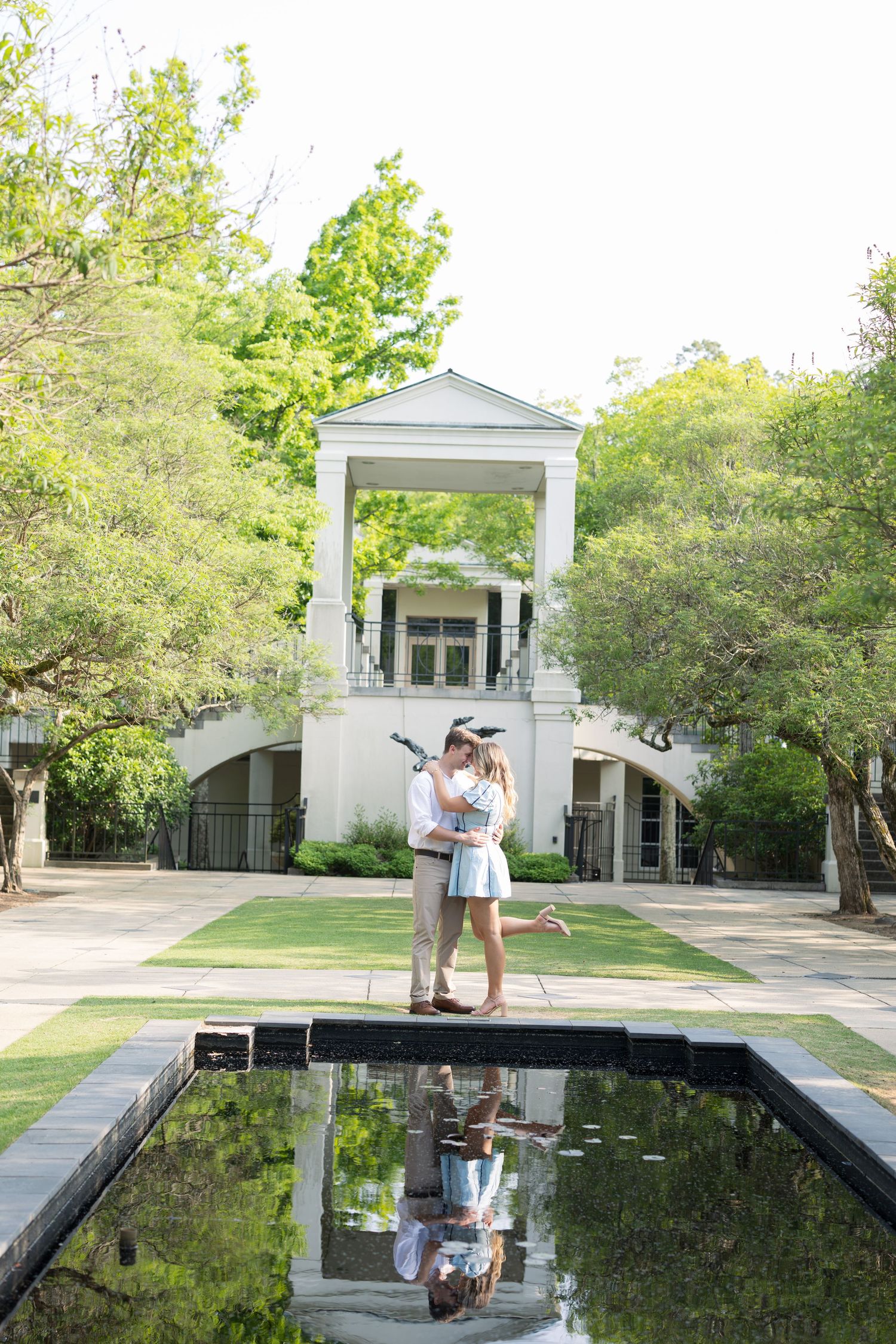 Couple embracing by a reflective pool in a lush garden with a white classical pavilion backdrop.